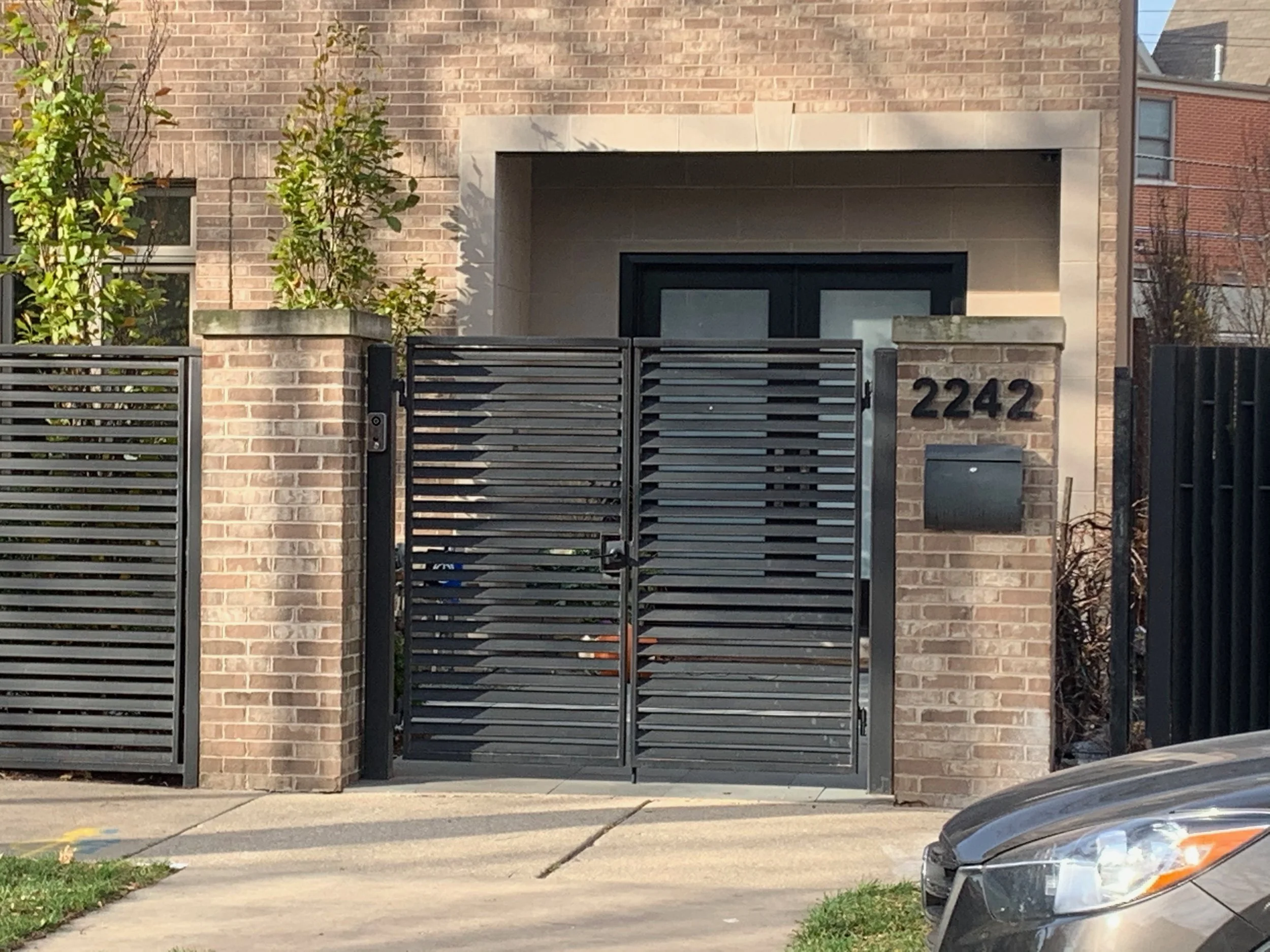Front view of a modern brick house with a black gate and the house number 2242 displayed on a brick pillar. There is a small tree and some plants on the left side, and part of a parked car is visible on the right.
