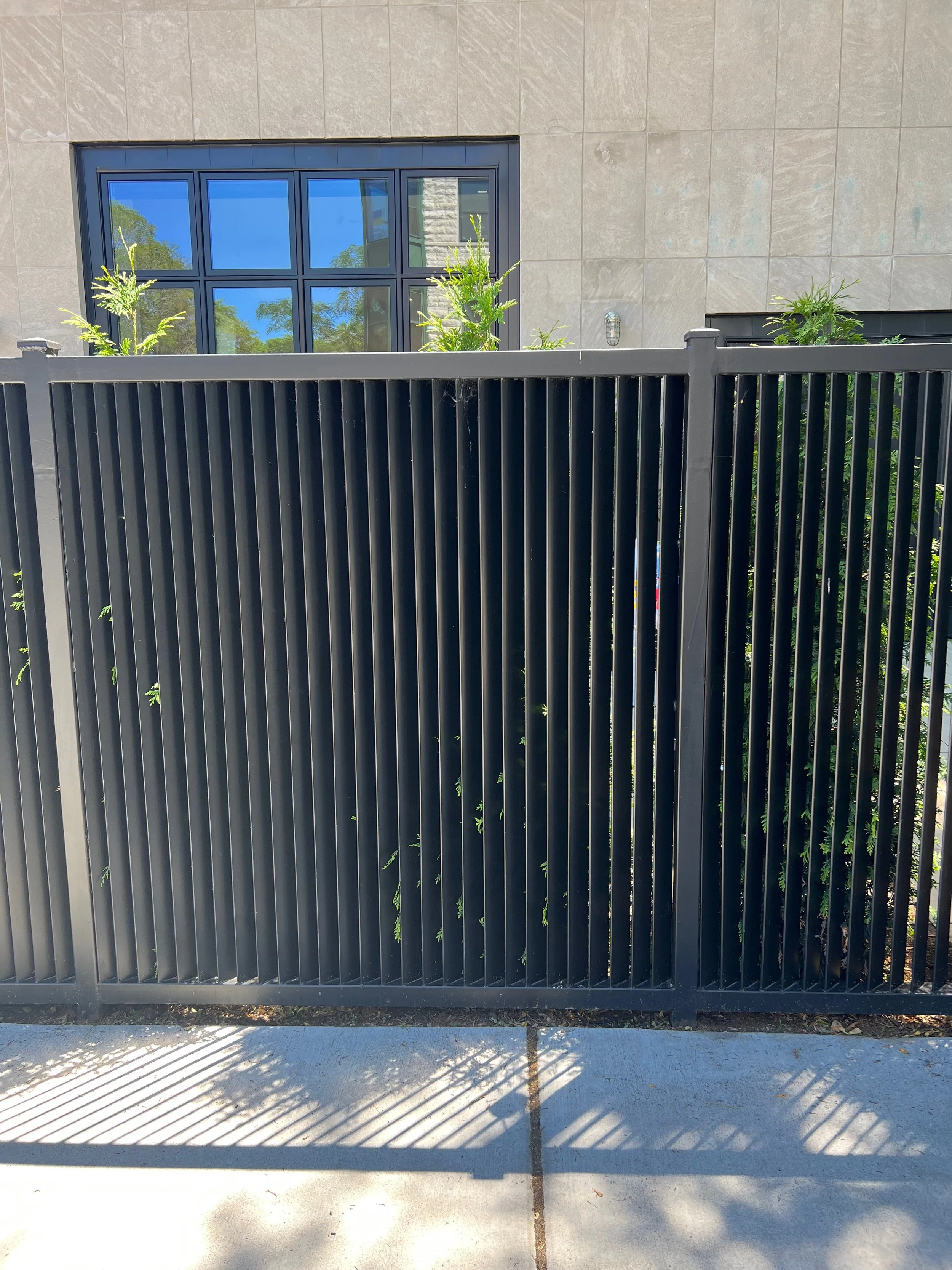 A black metal fence with vertical slats in front of a beige building wall, with a large window reflecting blue sky and trees, and a concrete sidewalk with shadows cast by the fence.