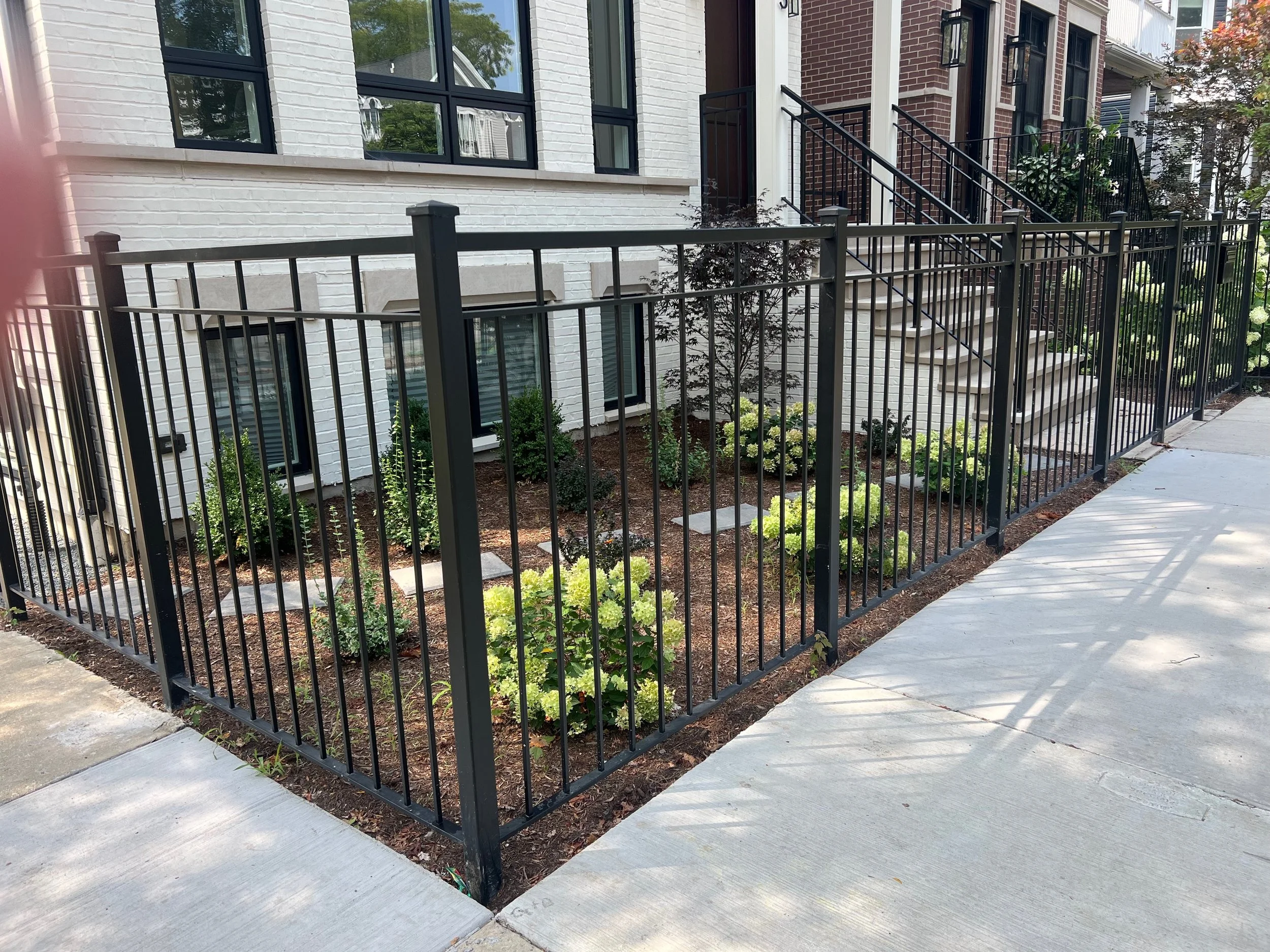 A black metal fence surrounds a small landscaped garden with green plants and a Japanese maple tree in front of a row of modern townhouses with brick and white painted exteriors, large windows, and exterior stairs.
