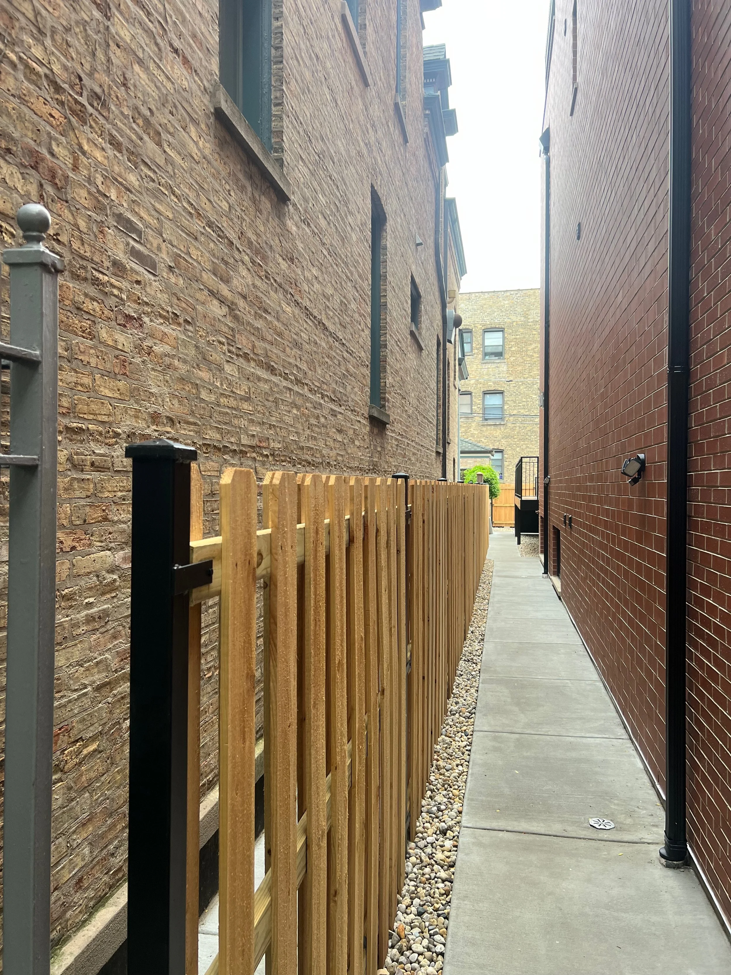 A narrow alley with a concrete sidewalk, a wooden fence on the left, brick buildings on both sides, and a potted plant in the distance.
