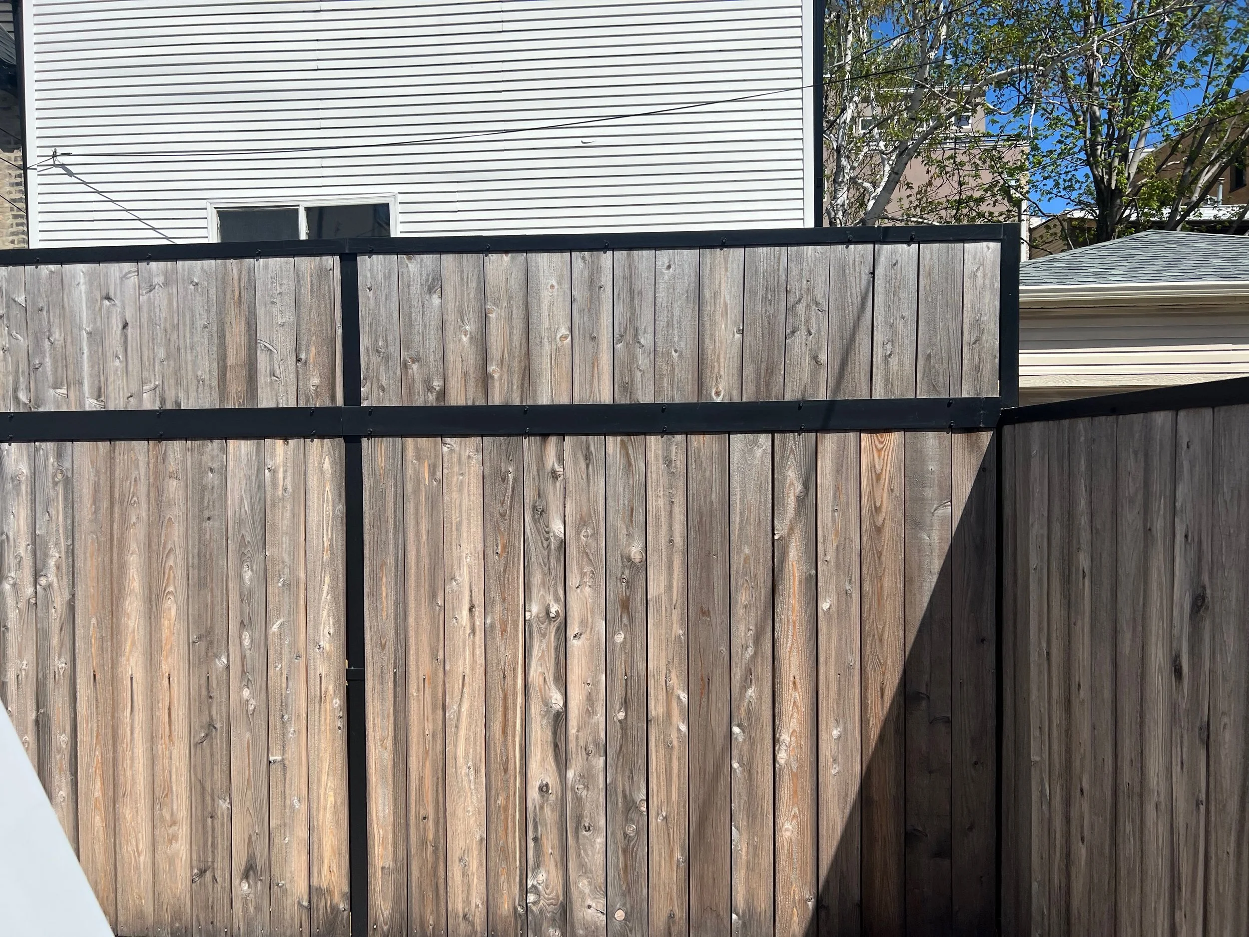 A wooden fence with a black metal frame, behind it a white house with horizontal siding, a window, and leafless trees under a clear blue sky.