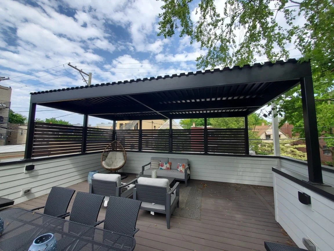 Outdoor rooftop patio with seating area, including chairs, a sofa with pillows, a hanging chair, and a table, surrounded by a black railing and partially covered by a metal roof, with trees and buildings in the background under a partly cloudy sky.