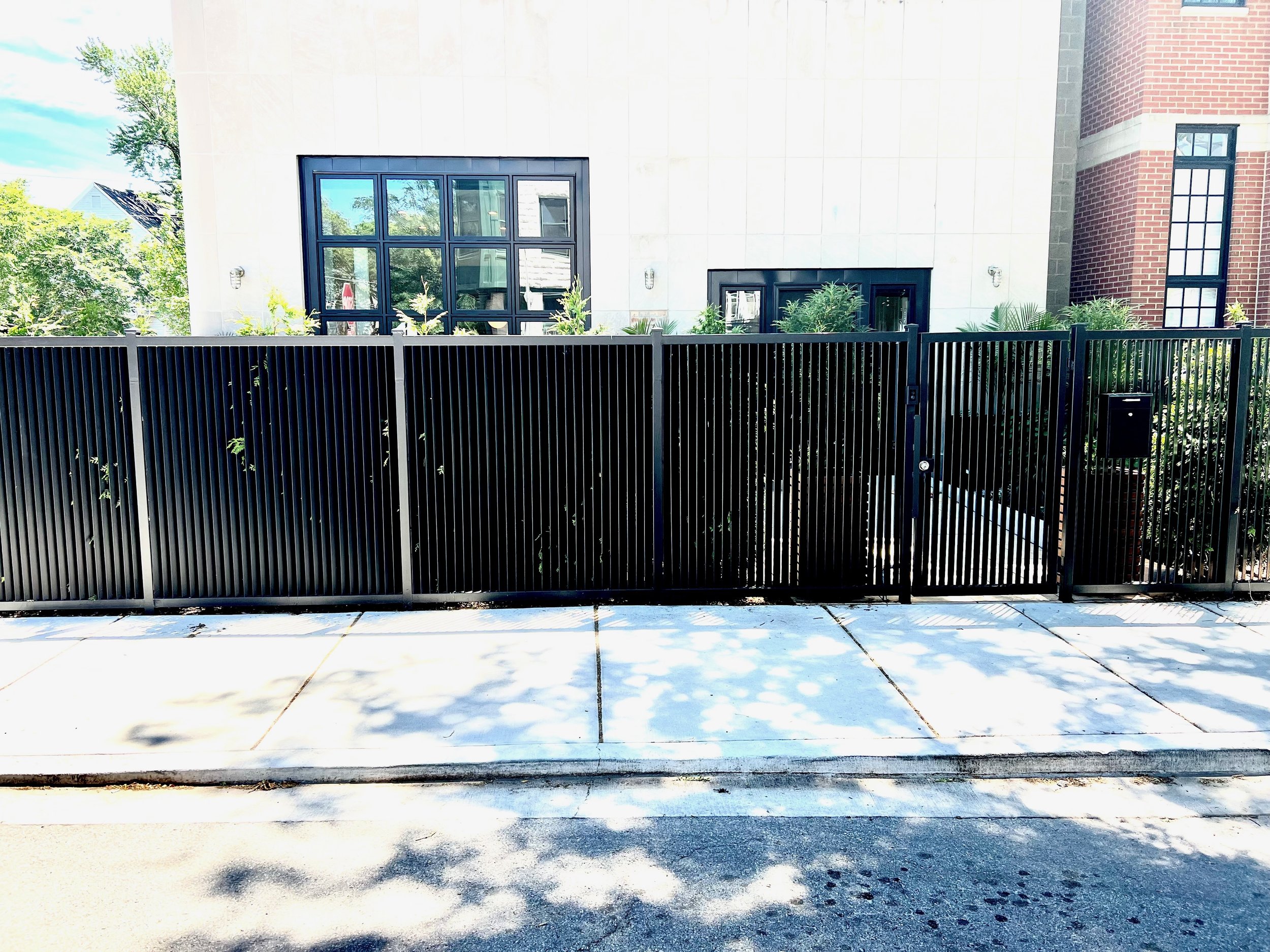 A black metal fence in front of a building with a large window and brick wall, with trees and a blue sky reflected in the window.
