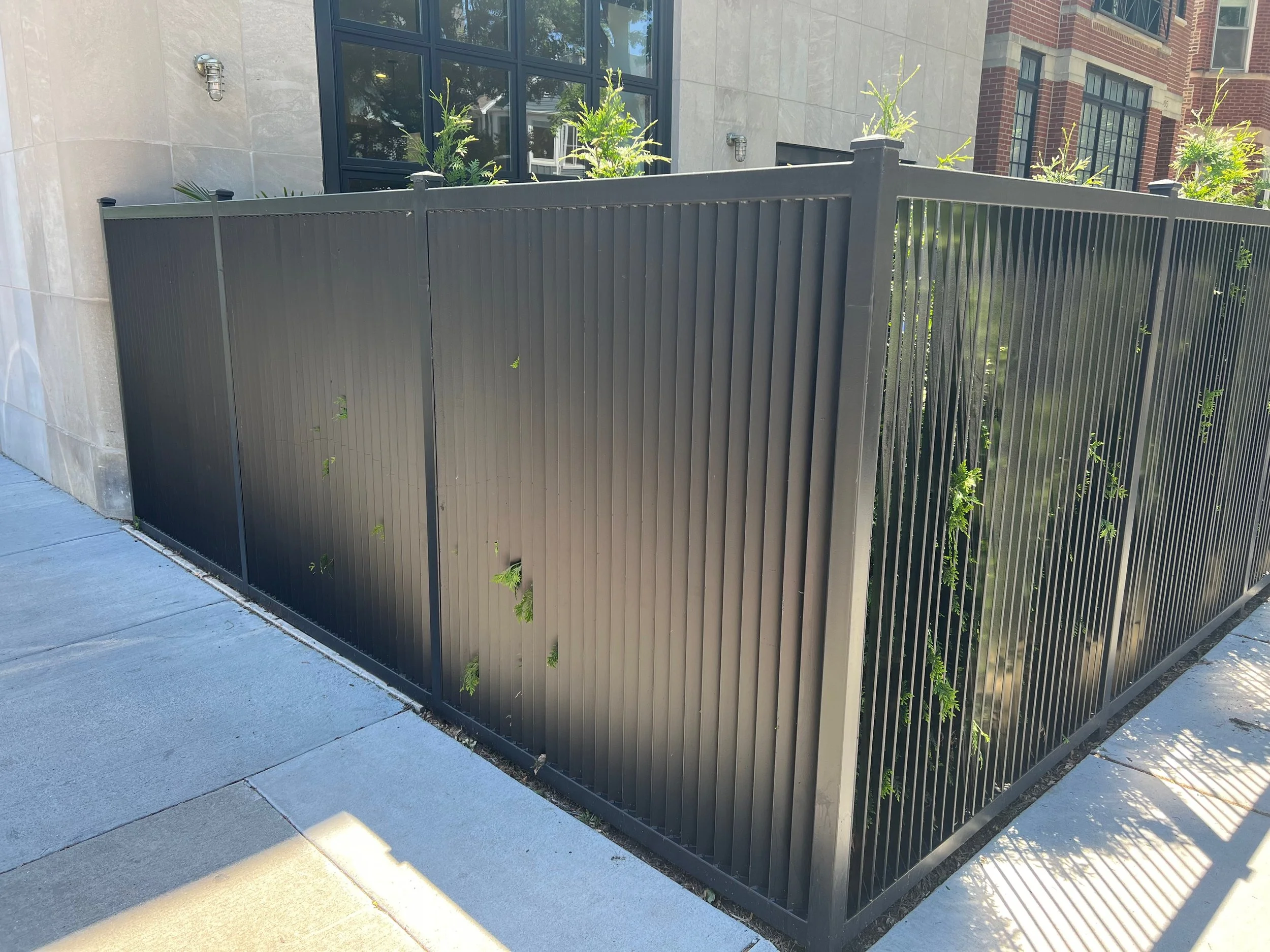 A black metal privacy fence surrounds a small garden with green plants. Behind the fence are the exterior walls of a modern building with large windows and brick accents.