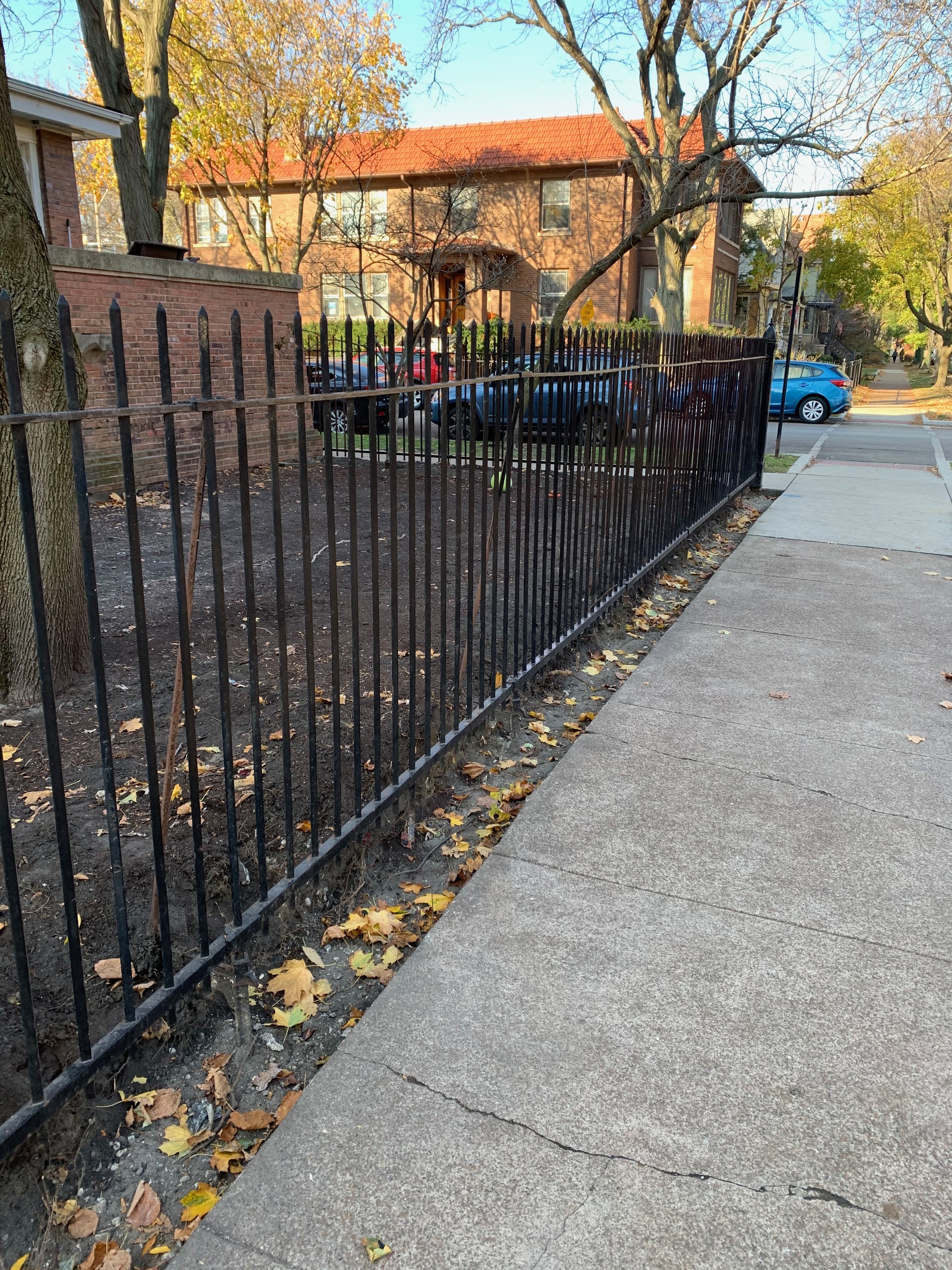 This image shows a sidewalk and a black iron fence with a small patches of soil and fallen yellow leaves on the ground. Behind the fence, there are trees with some yellow leaves, and a brick building with windows and a red tiled roof. Several parked 