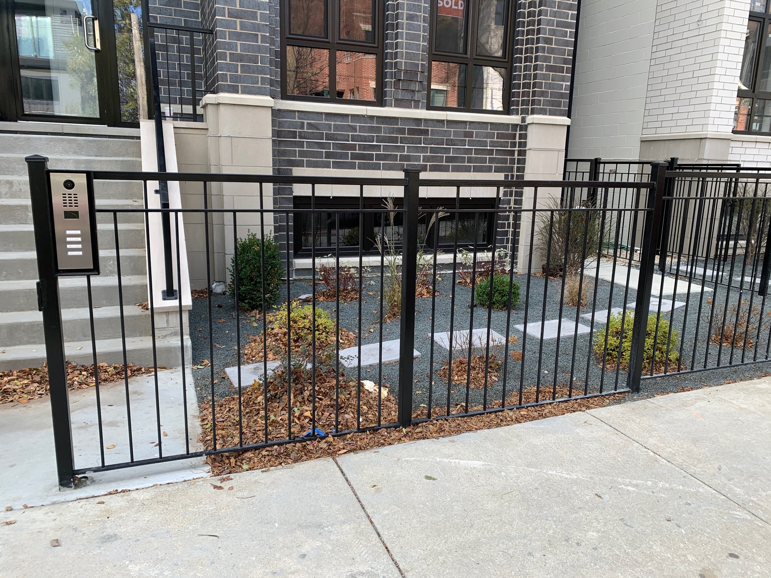 Front of a modern brick apartment building with stairs, a black metal fence, plant beds with bushes and dried leaves on the ground.