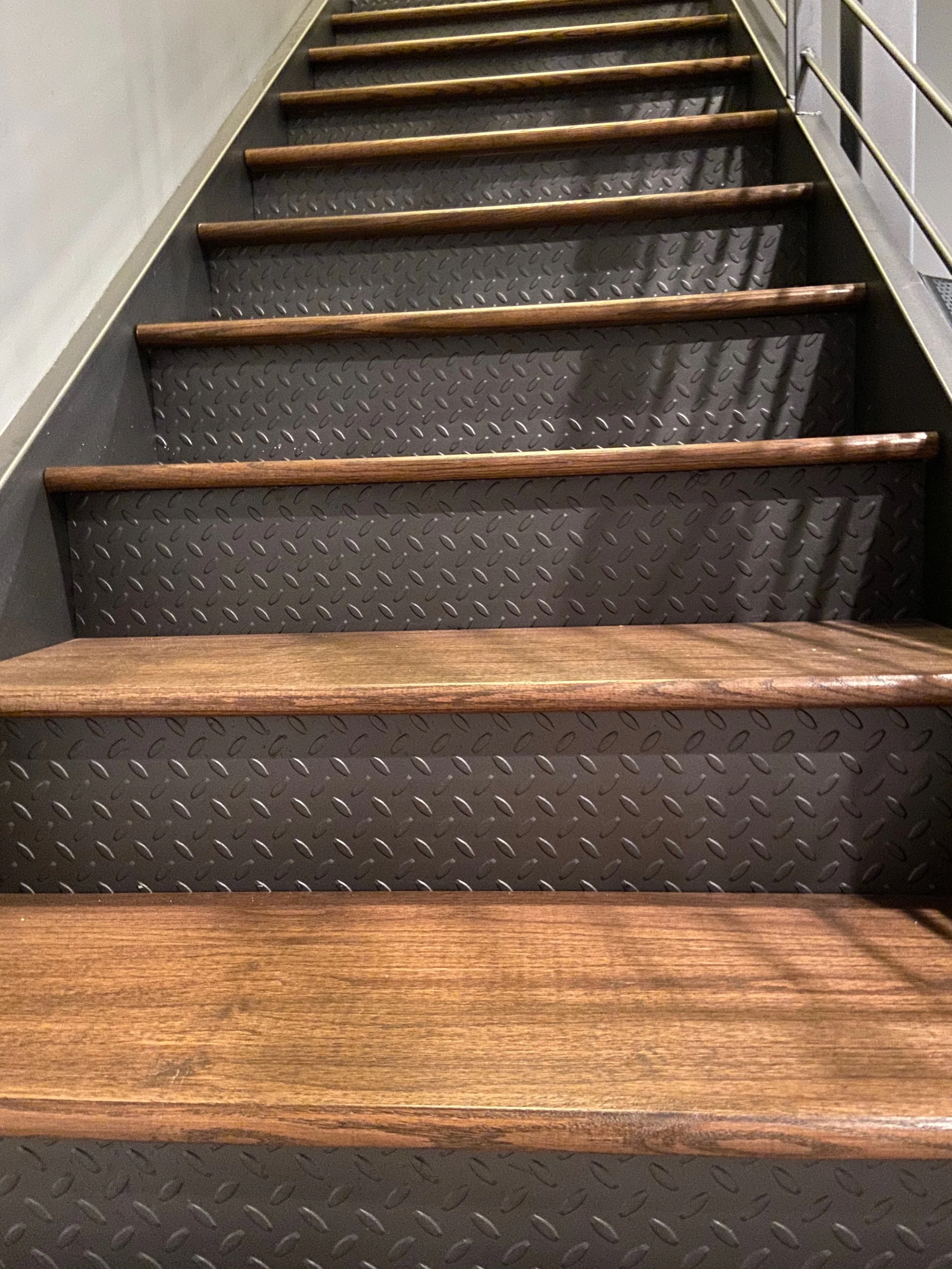 A staircase with dark metal steps featuring a diamond plate pattern, wooden handrails, and metal side panels with a similar diamond plate pattern.