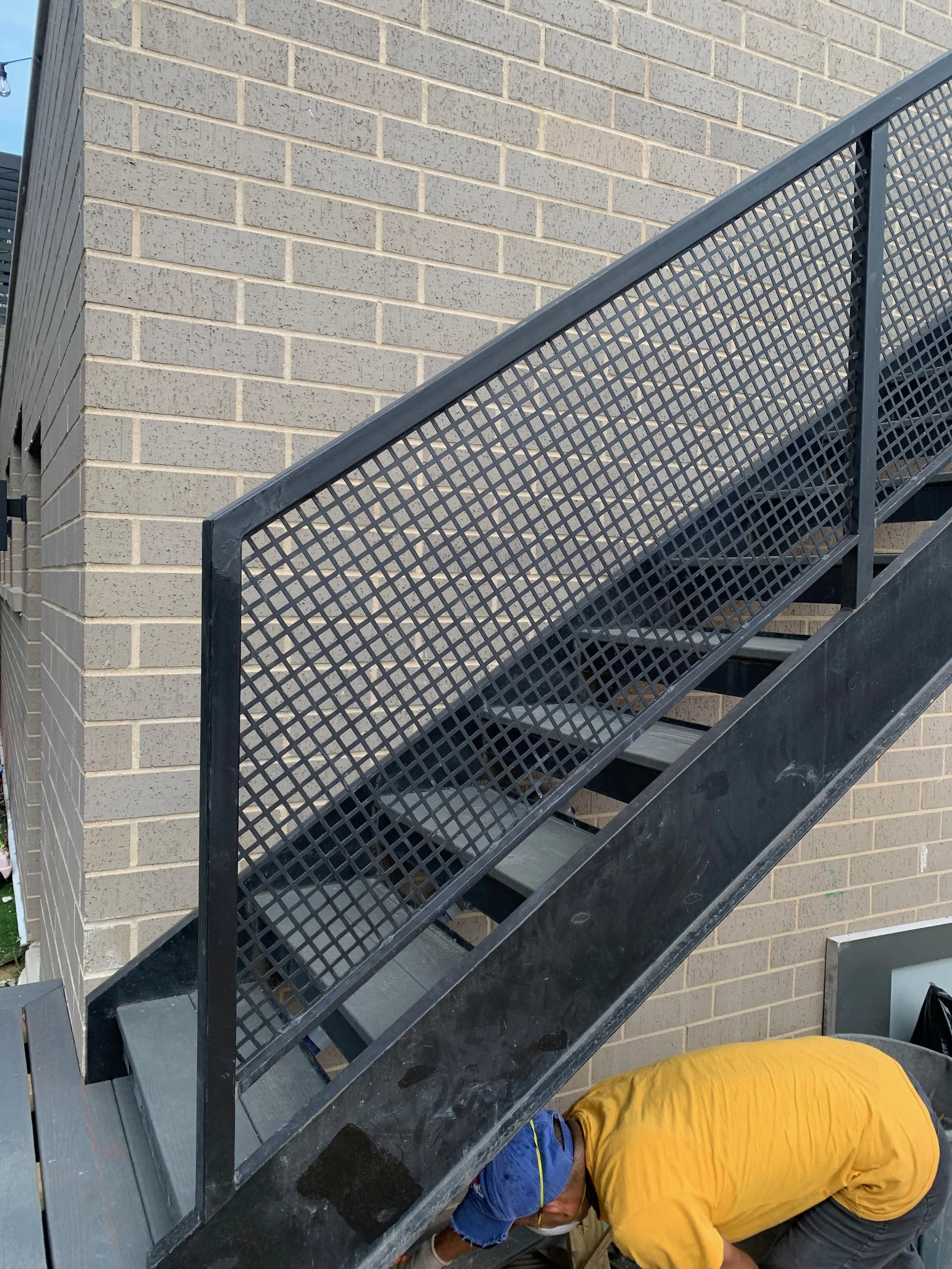 Two workers under a staircase, one in a yellow shirt and blue hat, the other in a blue shirt, working on a task near the building's brick wall.