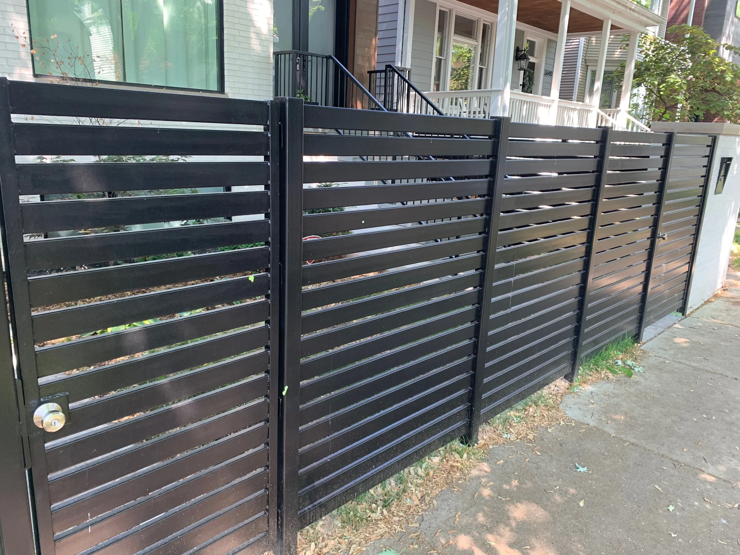 Black metal sliding gate closed in front of a sidewalk and residential houses with porches.
