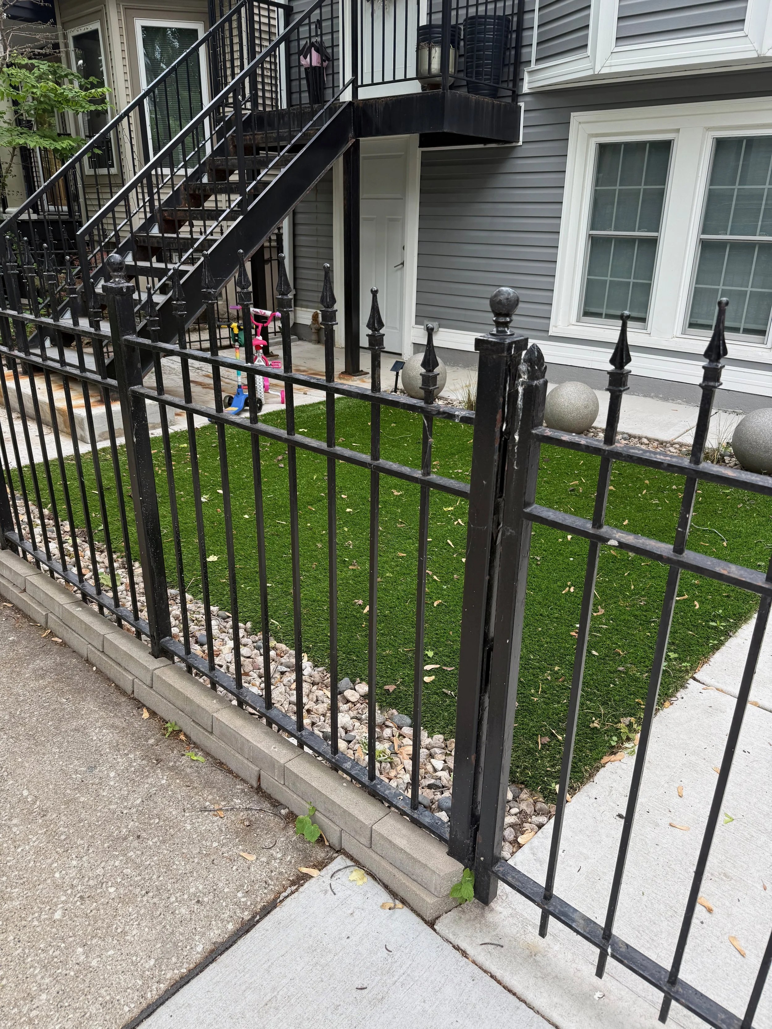 View of a residential backyard with a black metal fence, concrete pathway, small patch of artificial grass, and a gray house exterior with large windows and a staircase leading to a balcony.