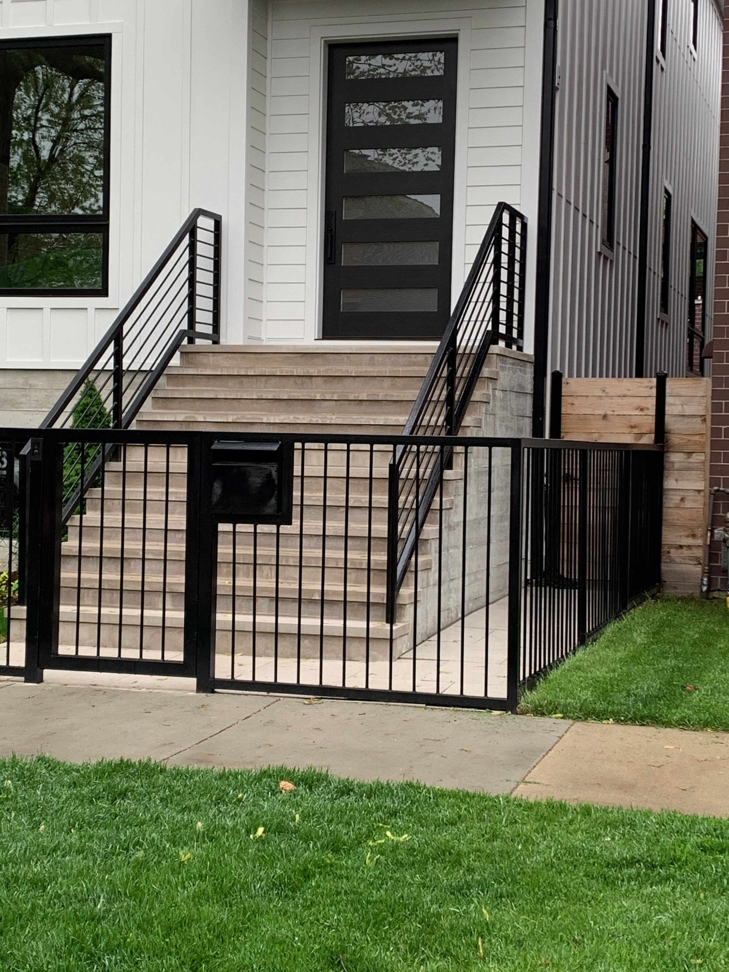 Front porch of a modern house with a black door, set of stairs, black metal railing, and a small fenced yard with green grass.