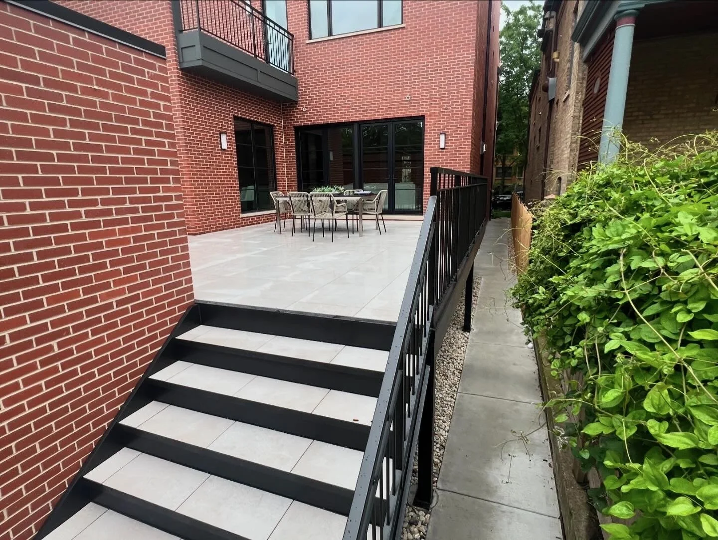 A modern outdoor patio with a dining table and chairs, accessed by a short set of black stairs, adjacent to a red brick building with sliding glass doors and a small balcony above. There are green bushes along the side.