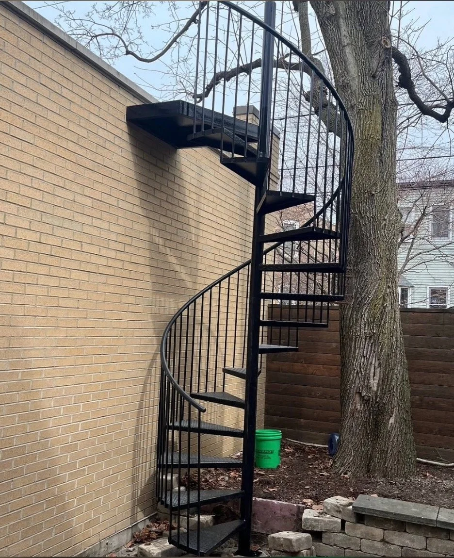 Black metal spiral staircase outdoors next to a brick wall and large tree.