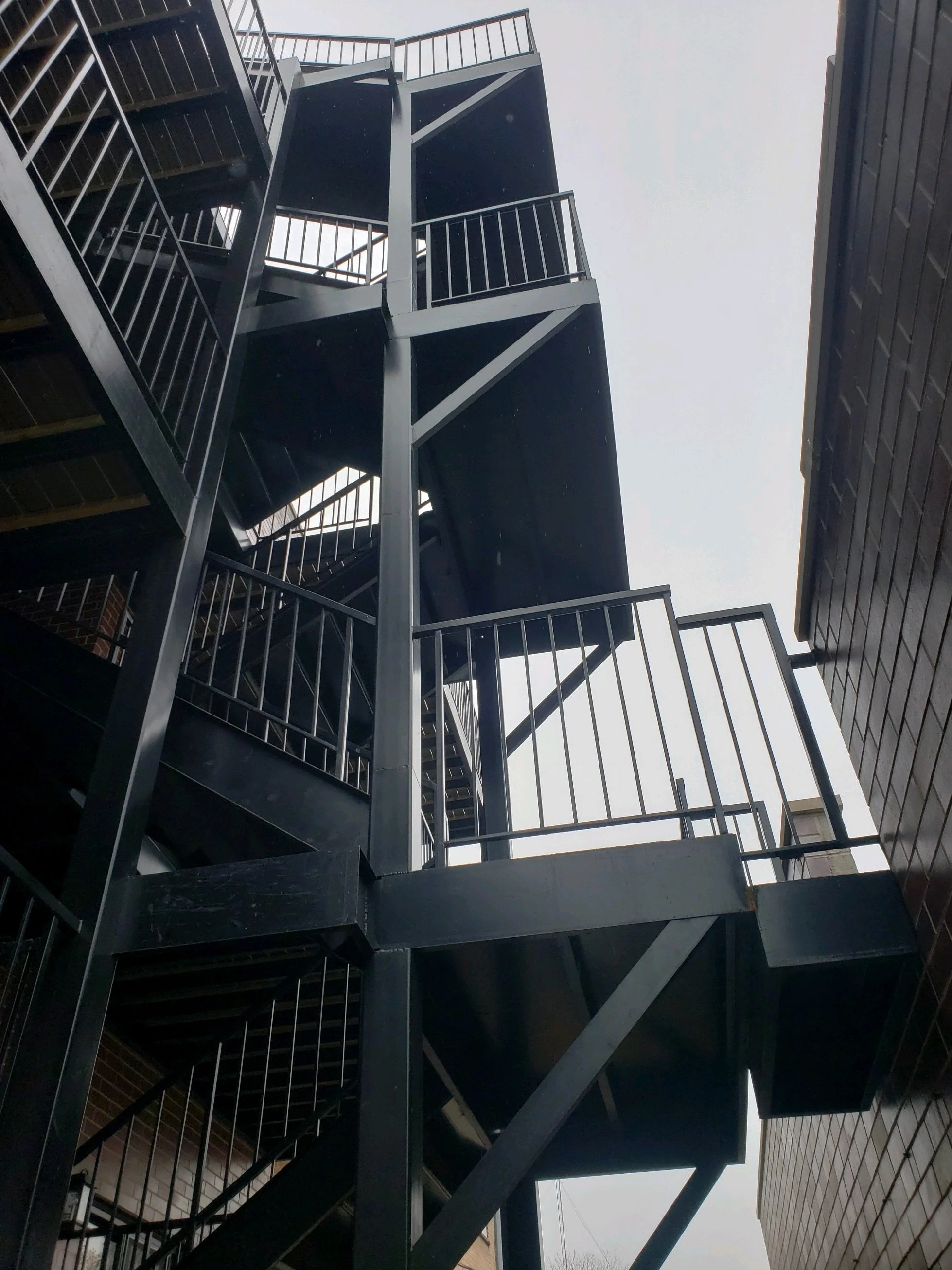 A black metal outdoor fire escape attached to the side of a brick building, with multiple staircases and landings, and metal railings, viewed from below against a cloudy sky.