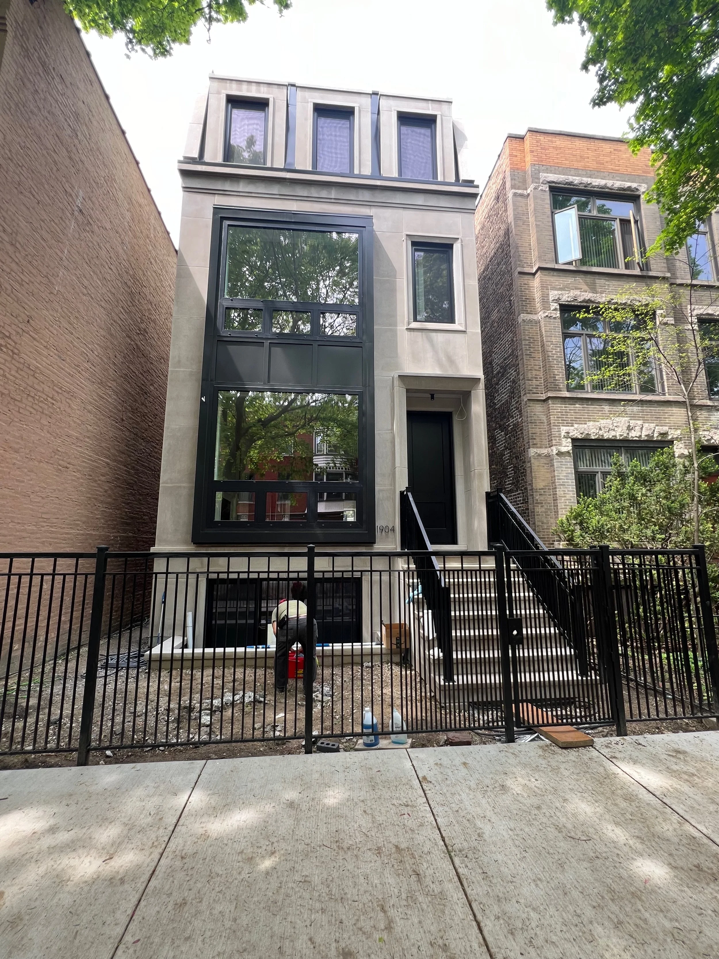 Front view of a modern three-story house under construction, with workers and construction tools in the yard, a black metal fence in the foreground, and neighboring buildings on either side.