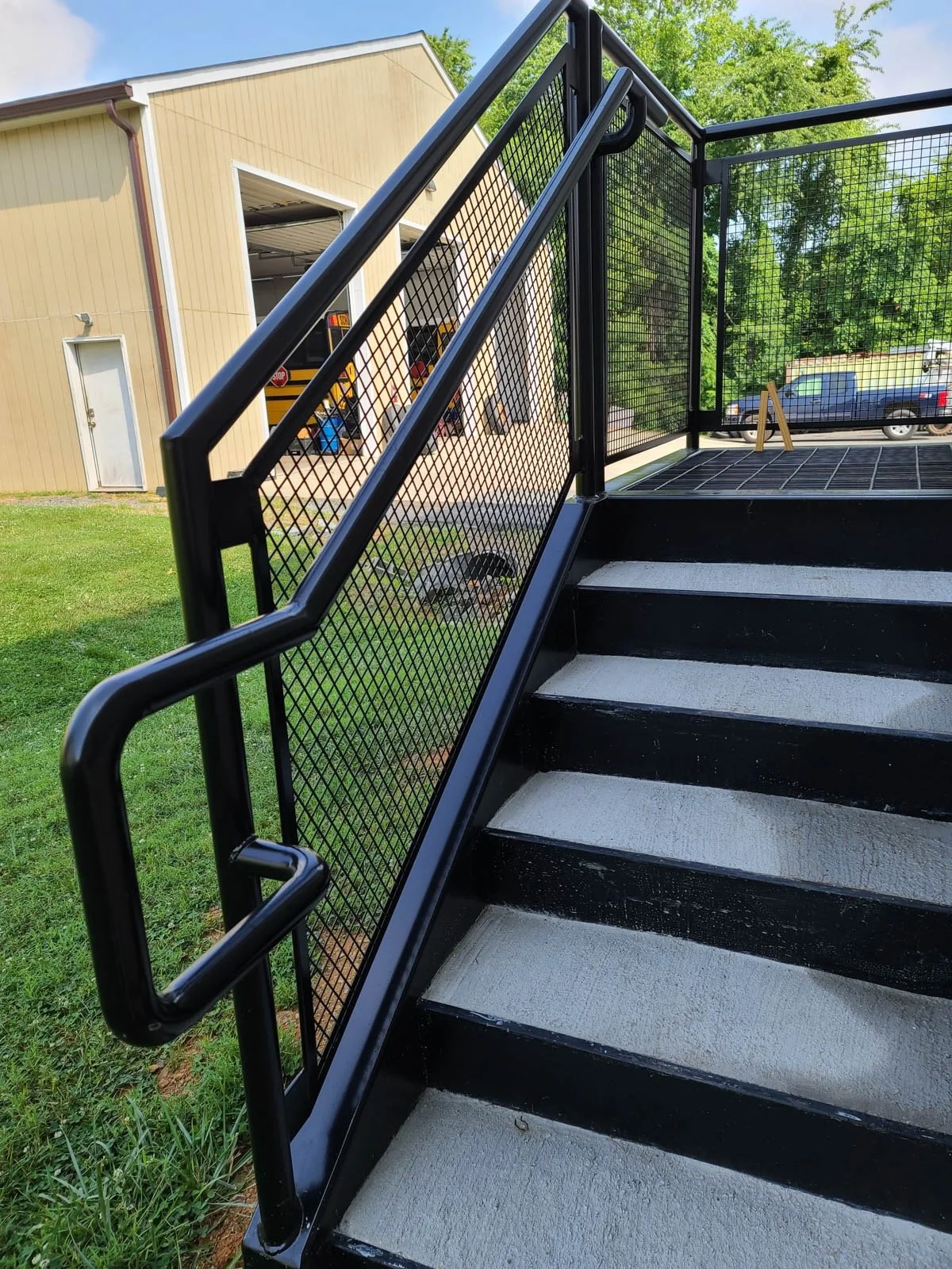 Black metal stairs with a textured white surface and a black mesh railing, outdoors on green grass with a beige building and parked cars in the background.