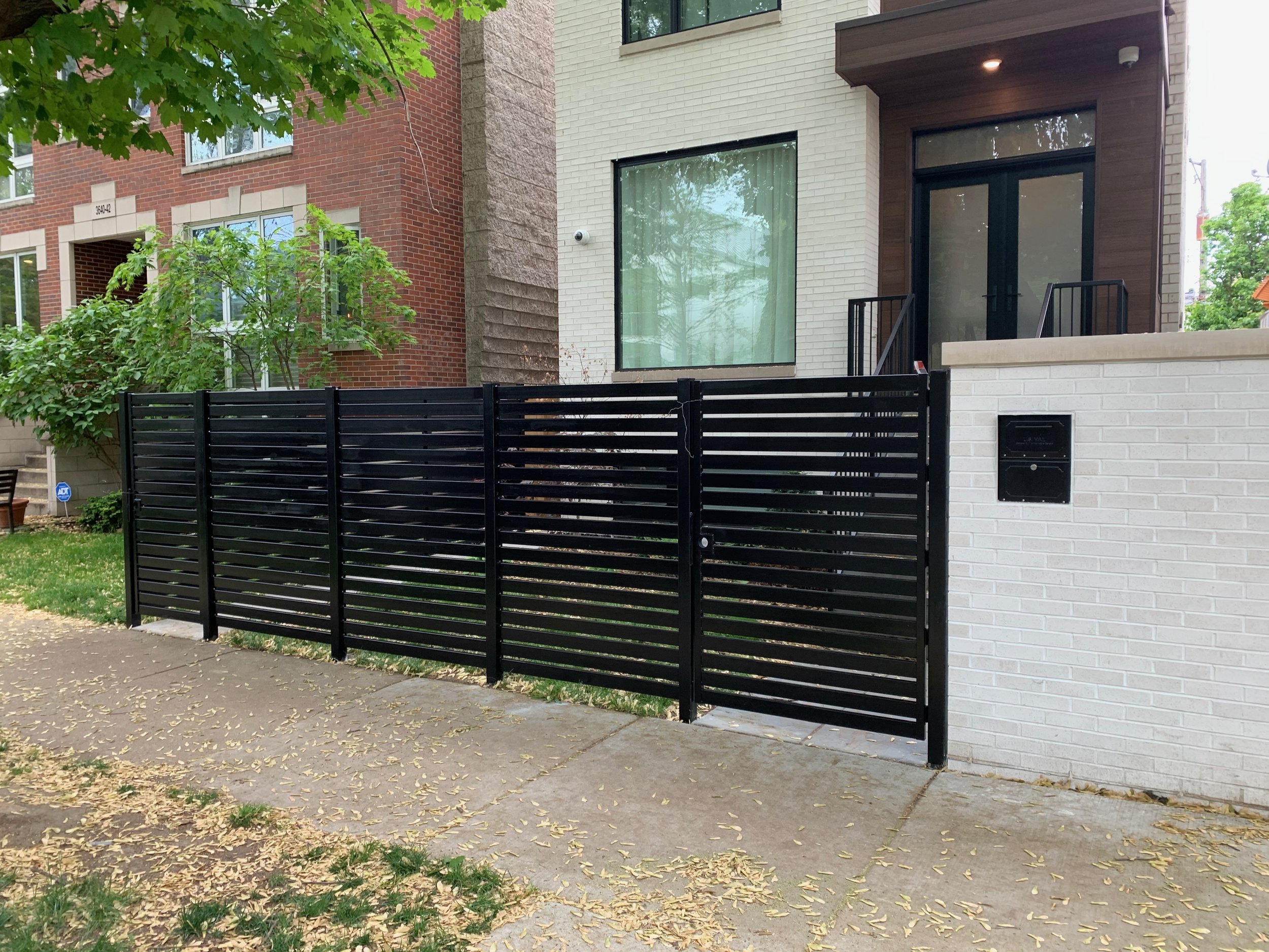 View of a modern residential building with a black metal gate and a white brick wall, trees and greenery around, sidewalk with fallen leaves.