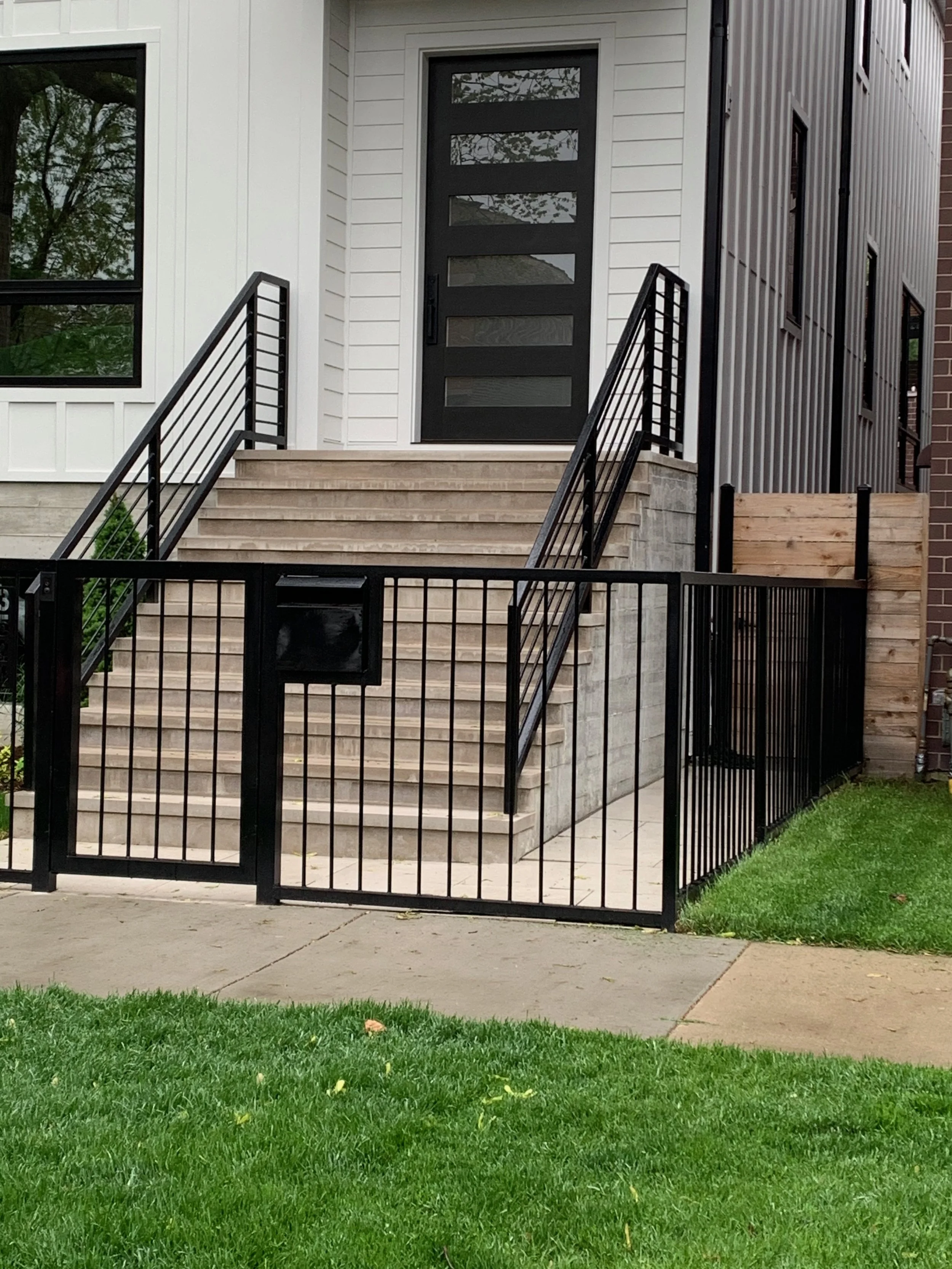 Front porch of a modern house with a black door, steps, and metal railing, surrounded by a small grass yard.