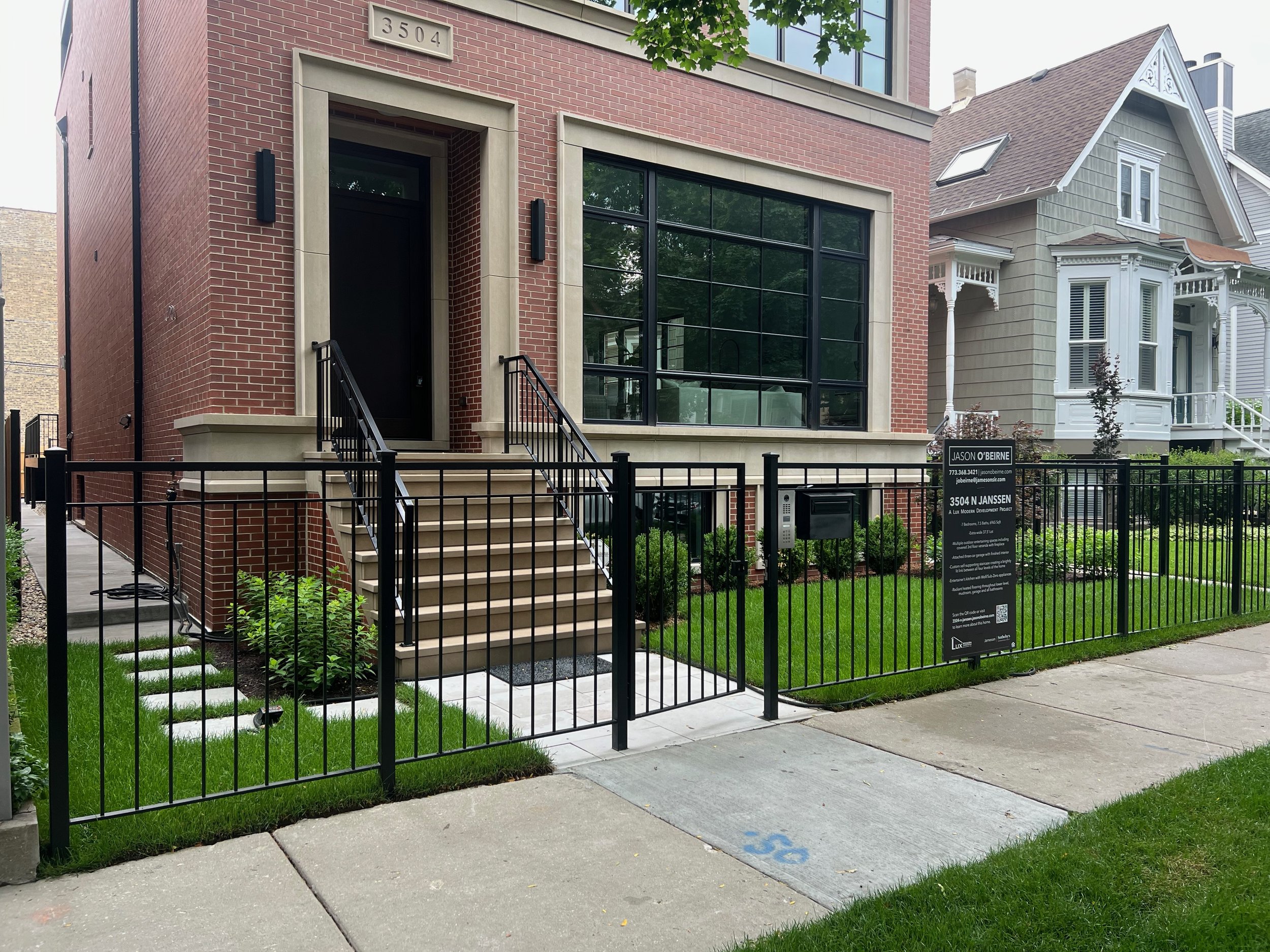 Modern brick and glass residential building with stairs, black fence, and a yard, located at 3504 N Janssen. Adjacent to an older Victorian-style house with white trim.