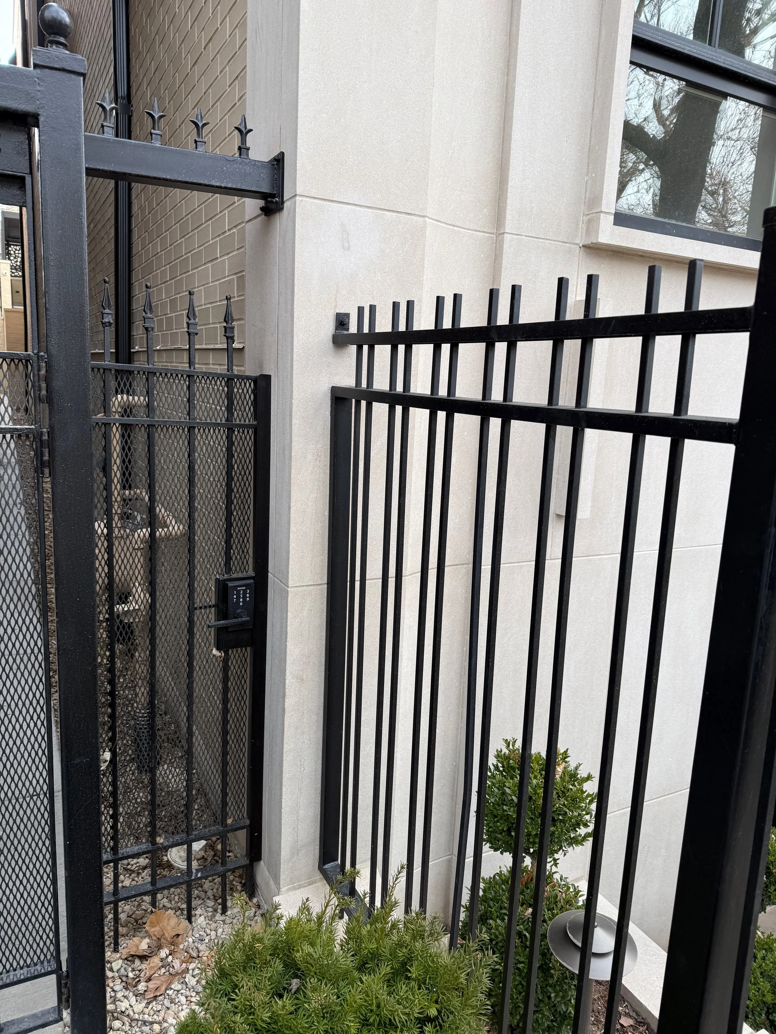 A black metal gate and fence in front of a beige building wall with a window, a small bush, outdoor solar light, and gravel on the ground.