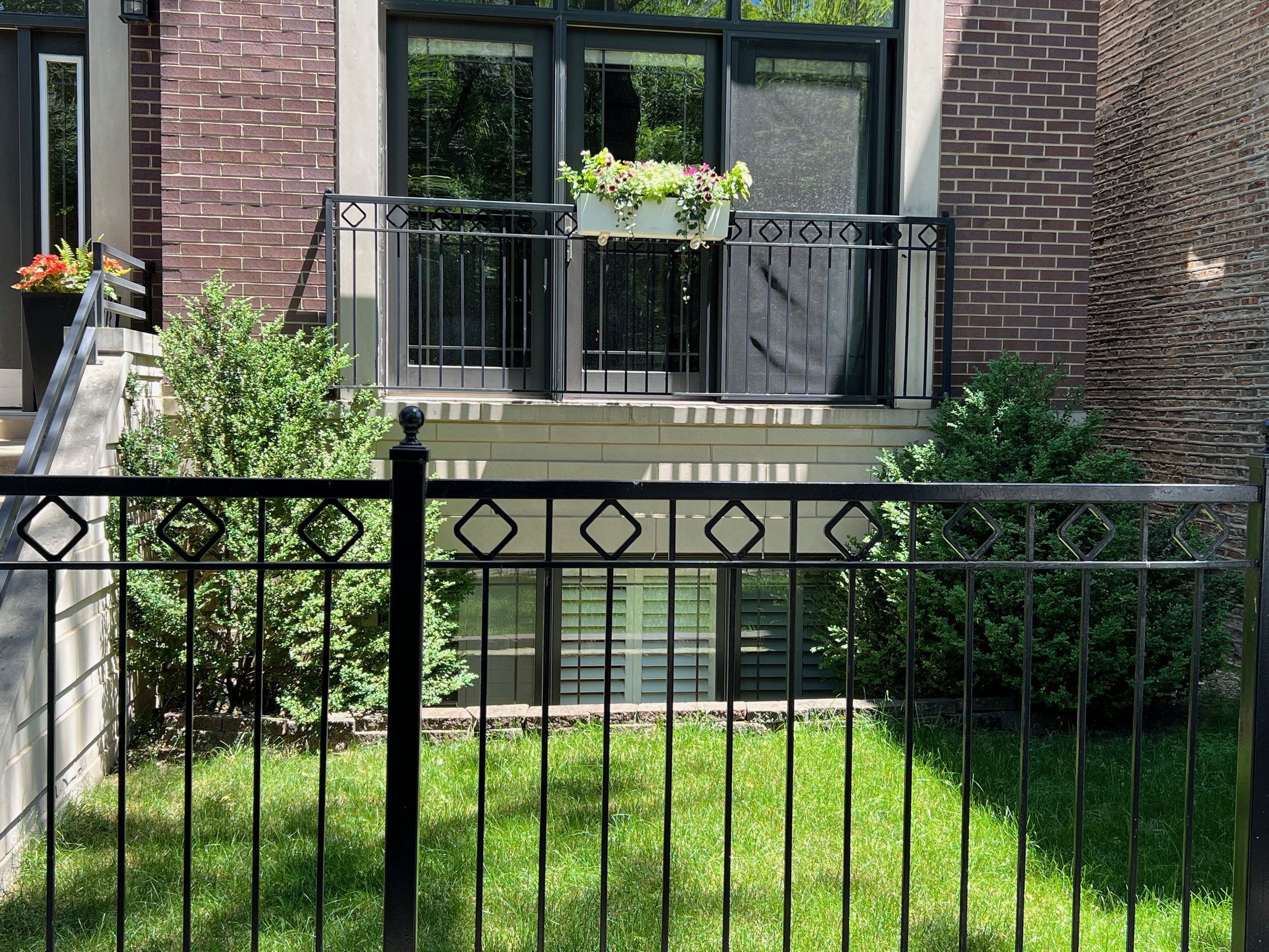 A small balcony with black metal railing and potted flowers, attached to a brick apartment building with large glass doors, surrounded by greenery and a grassy yard.