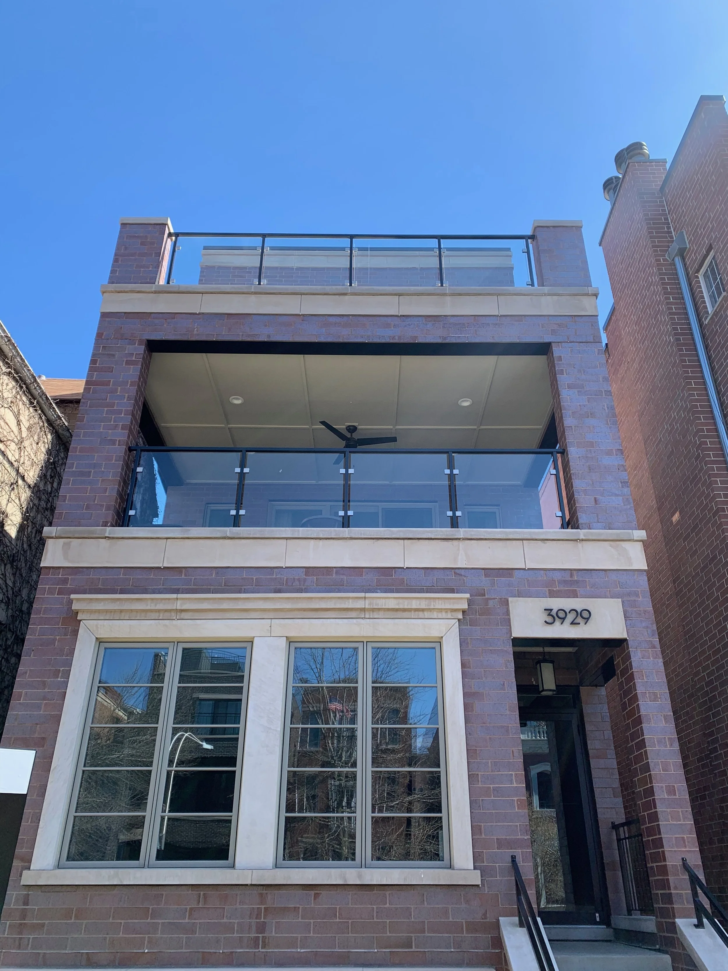 The image shows the front of a modern brick apartment building with a balcony on the top floor, featuring glass railings. The building has large white-framed windows on the lower level and the house number '3929' displayed near the entrance. There is