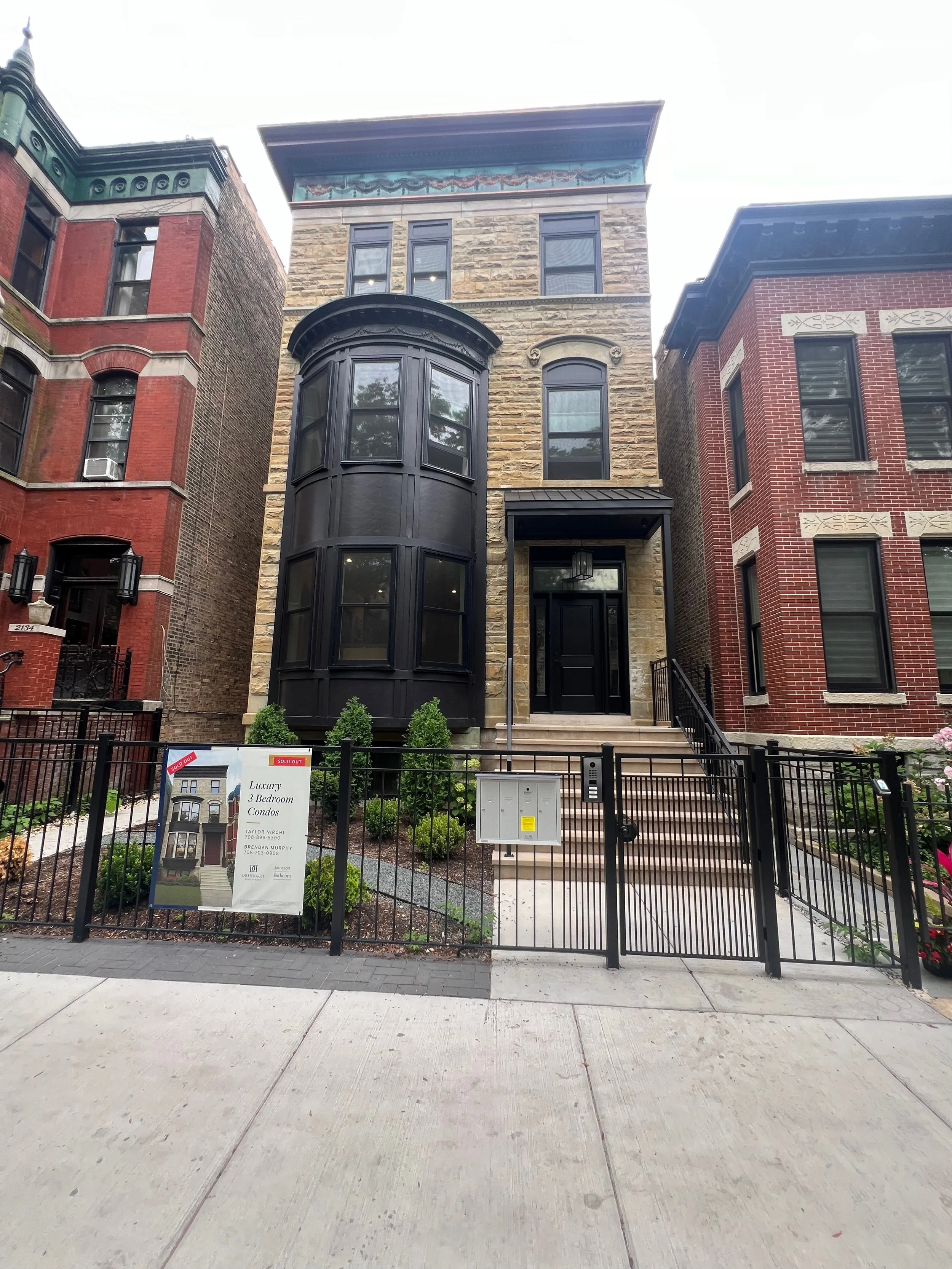 Front view of a three-story townhouse with a black bay window, beige brick exterior, and a small front yard enclosed by a black metal fence. There is a sign in the yard advertising luxury 3 bedroom condos.