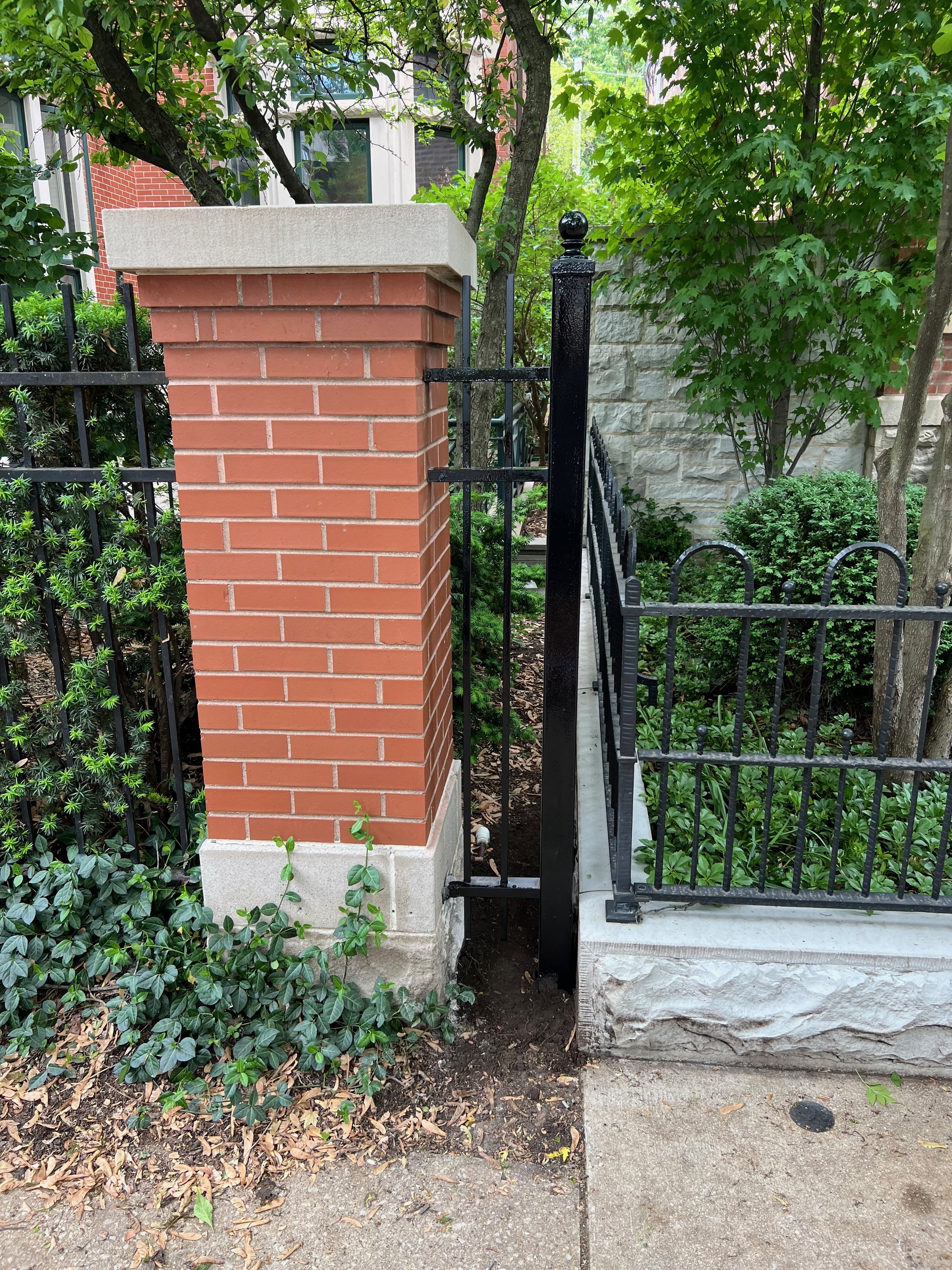A brick and stone pillar with a concrete cap, next to a black wrought iron fence and gate, surrounded by green bushes and trees in a residential area.