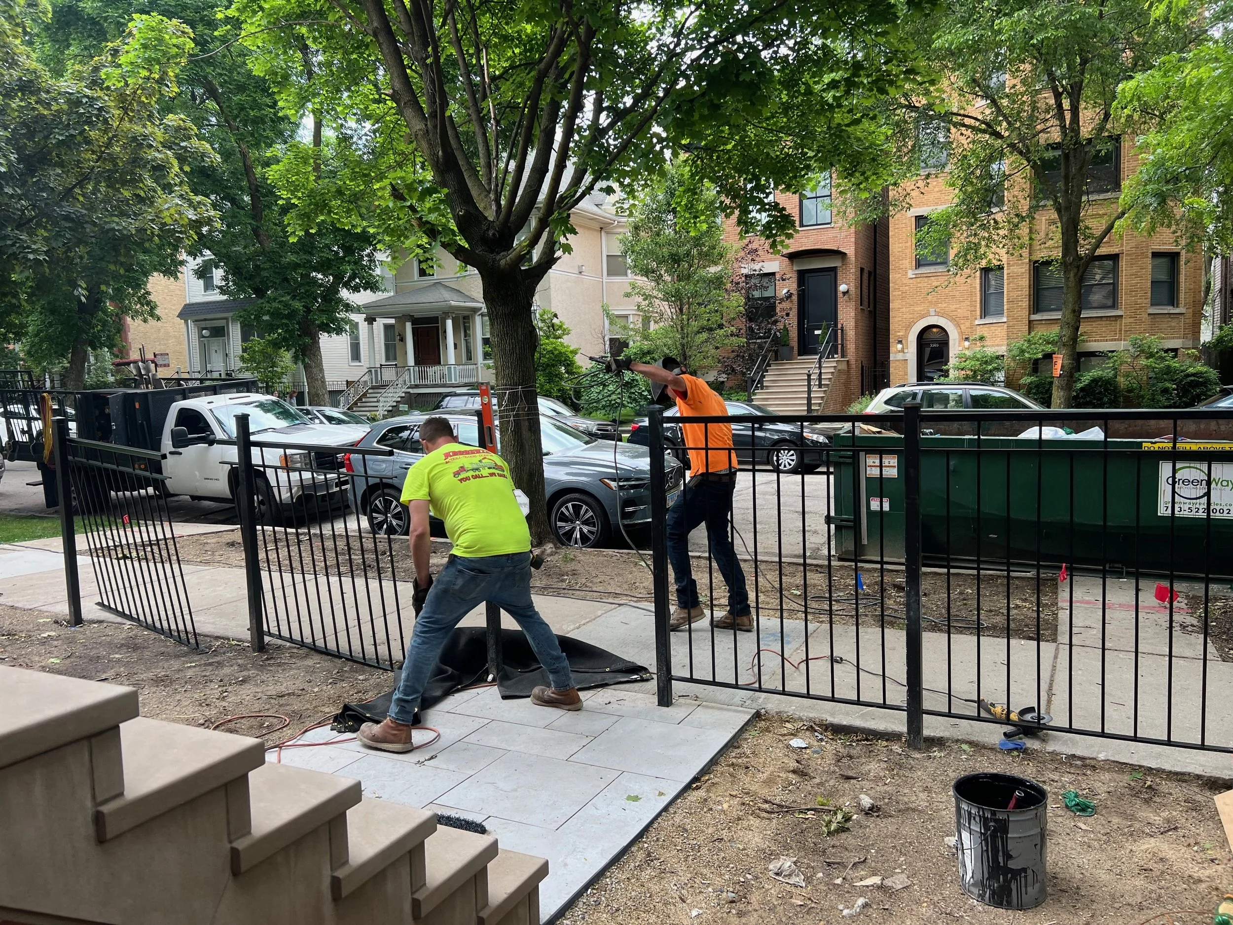 Two construction workers installing paving stones on a sidewalk in front of residential buildings with trees and parked cars.