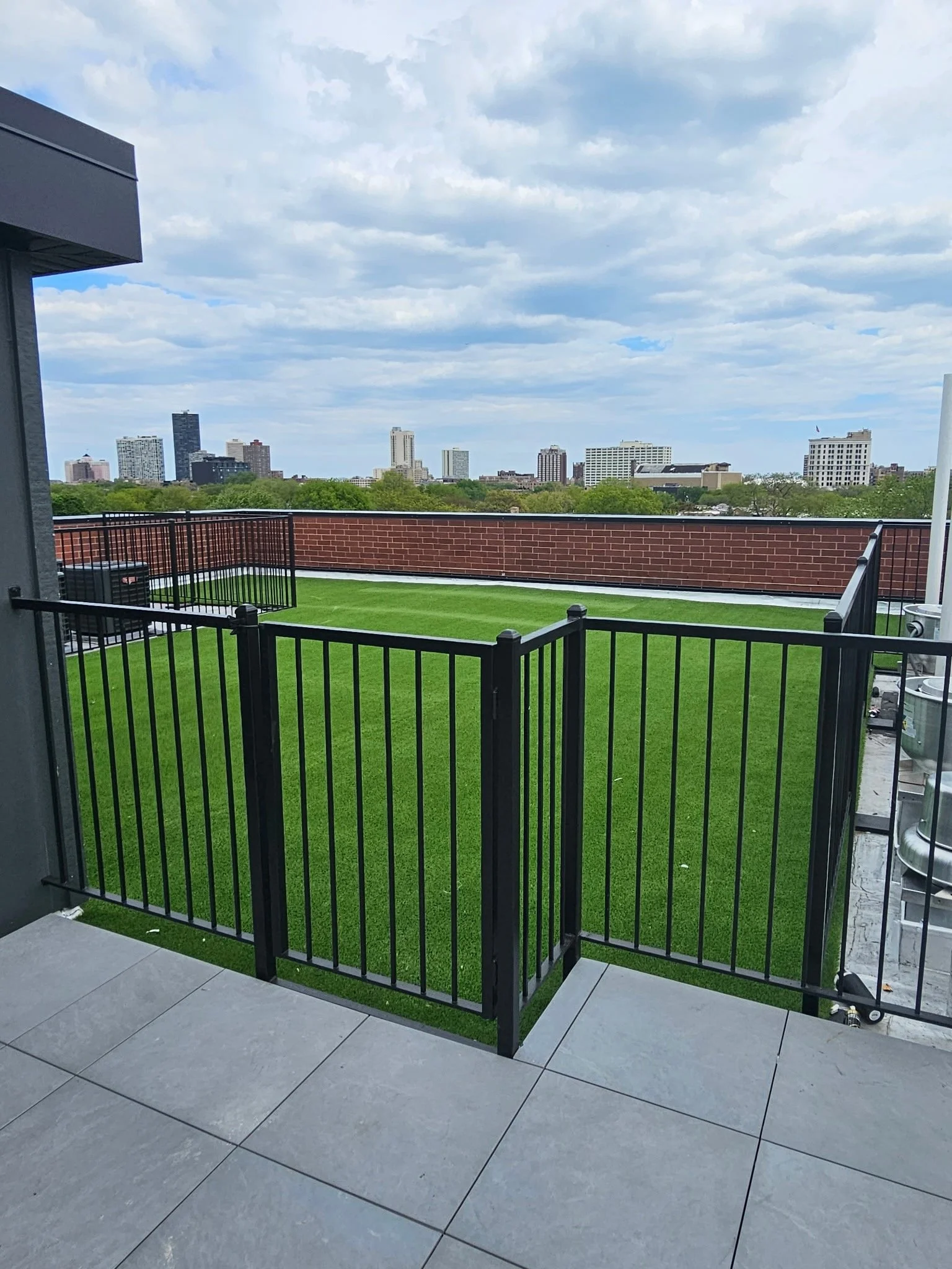 Balcony with artificial grass, black metal fence, and city skyline in the background under a partly cloudy sky.
