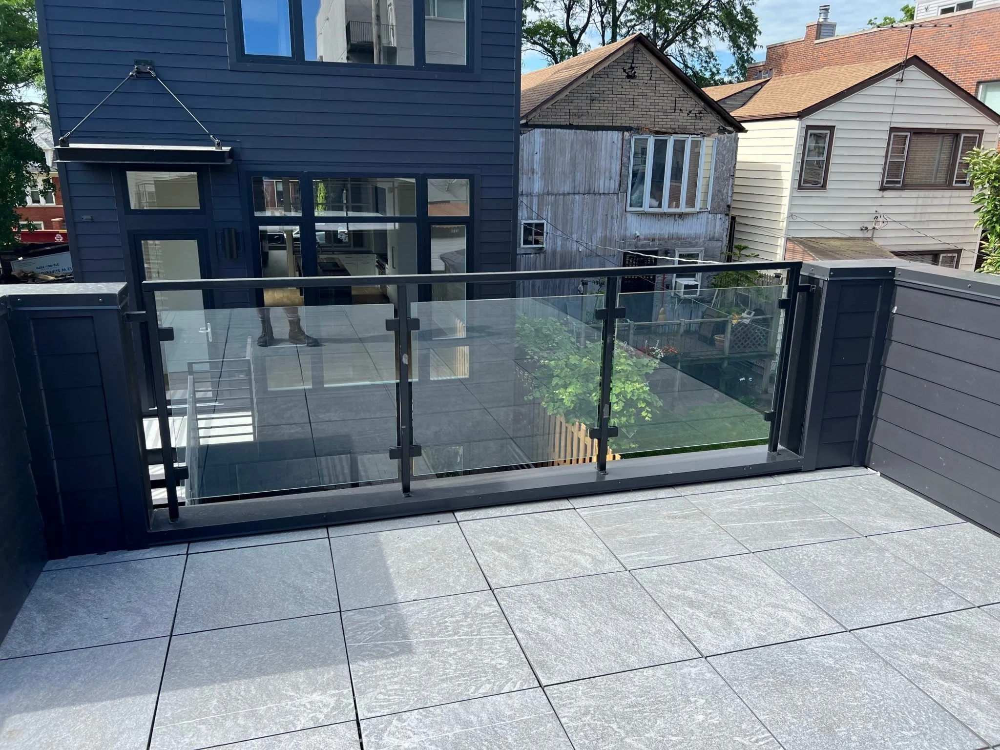 View from a balcony showing grey tiled flooring, a glass railing, and houses with different exterior siding styles in the background.