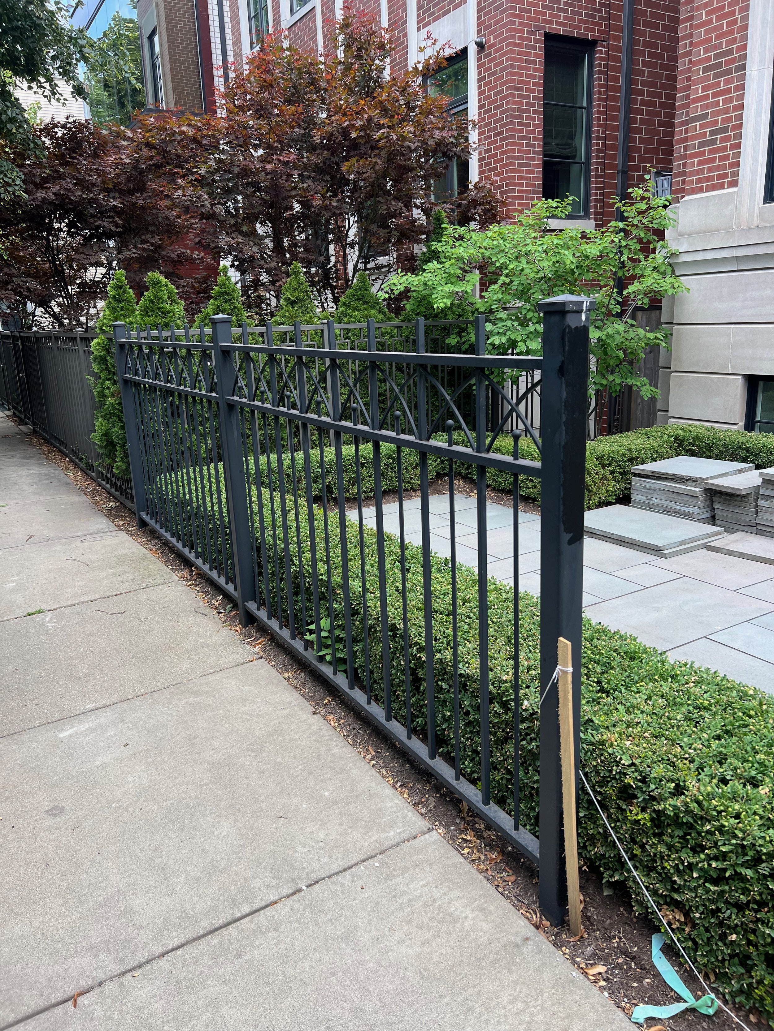 A black metal fence with decorative elements separates a sidewalk from a garden with various green plants and shrubbery in front of a brick residential building.