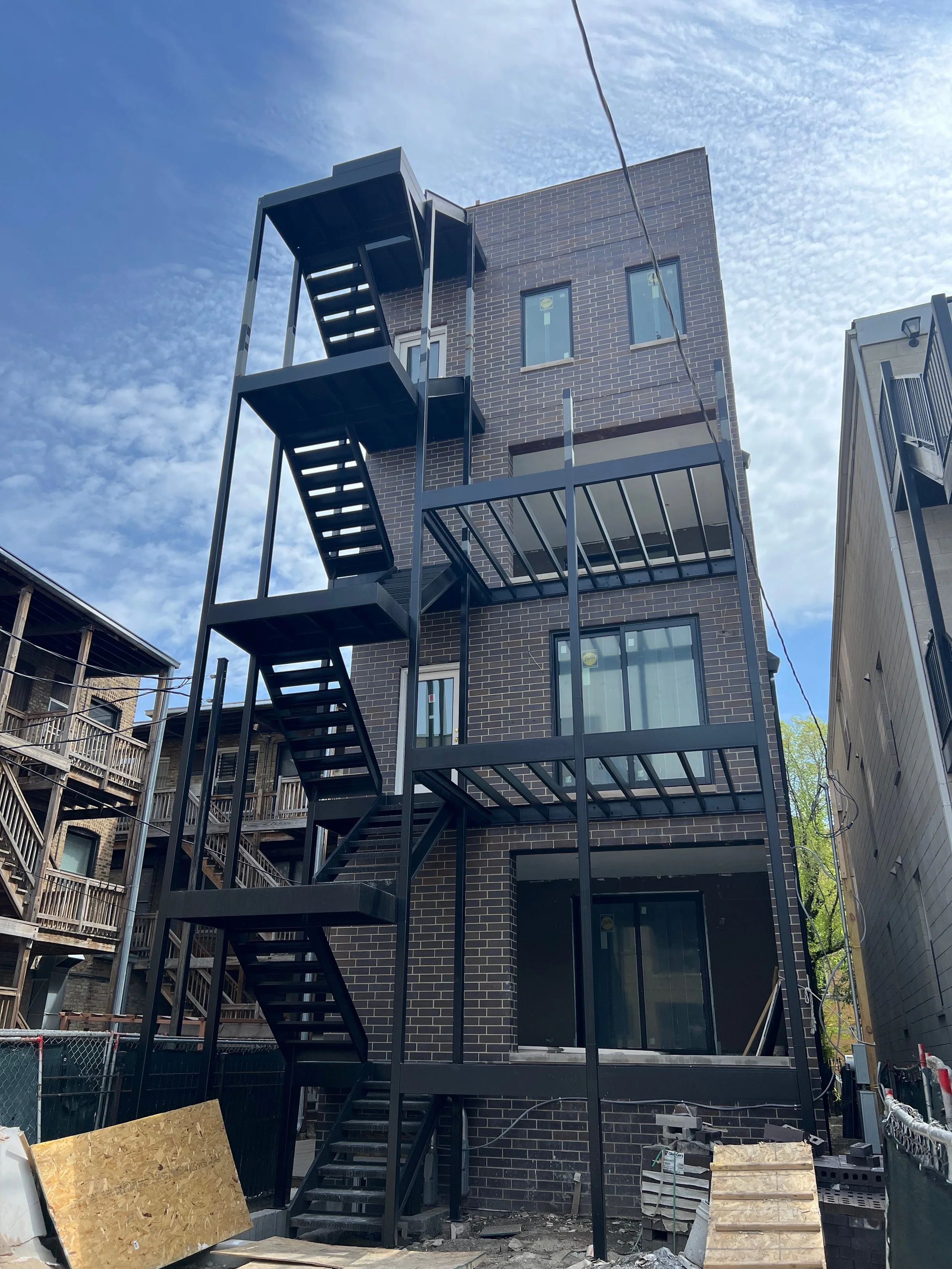 A staircase attached to the side of a multi-story brick building under construction. The staircase is made of black metal with open risers. There is construction materials and debris on the ground, with a plywood sheet leaning against a railing in th