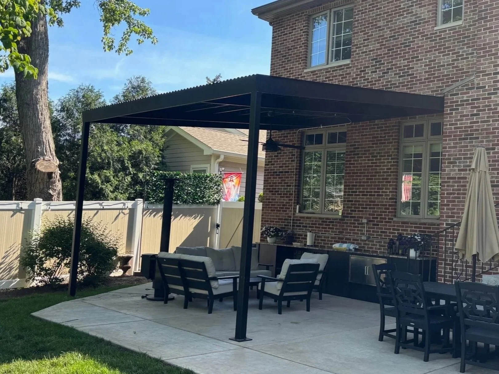 Backyard patio with seating area under a black pergola, brick house in the background, white fence, and green lawn.