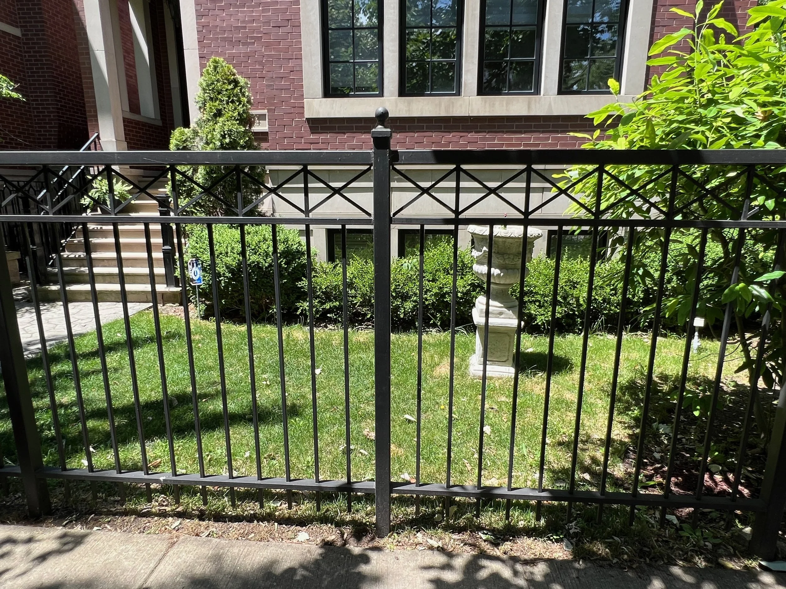View of a backyard garden with a black metal fence, green grass, shrubs, and a concrete bird bath or pedestal, in front of a brick building with large windows.