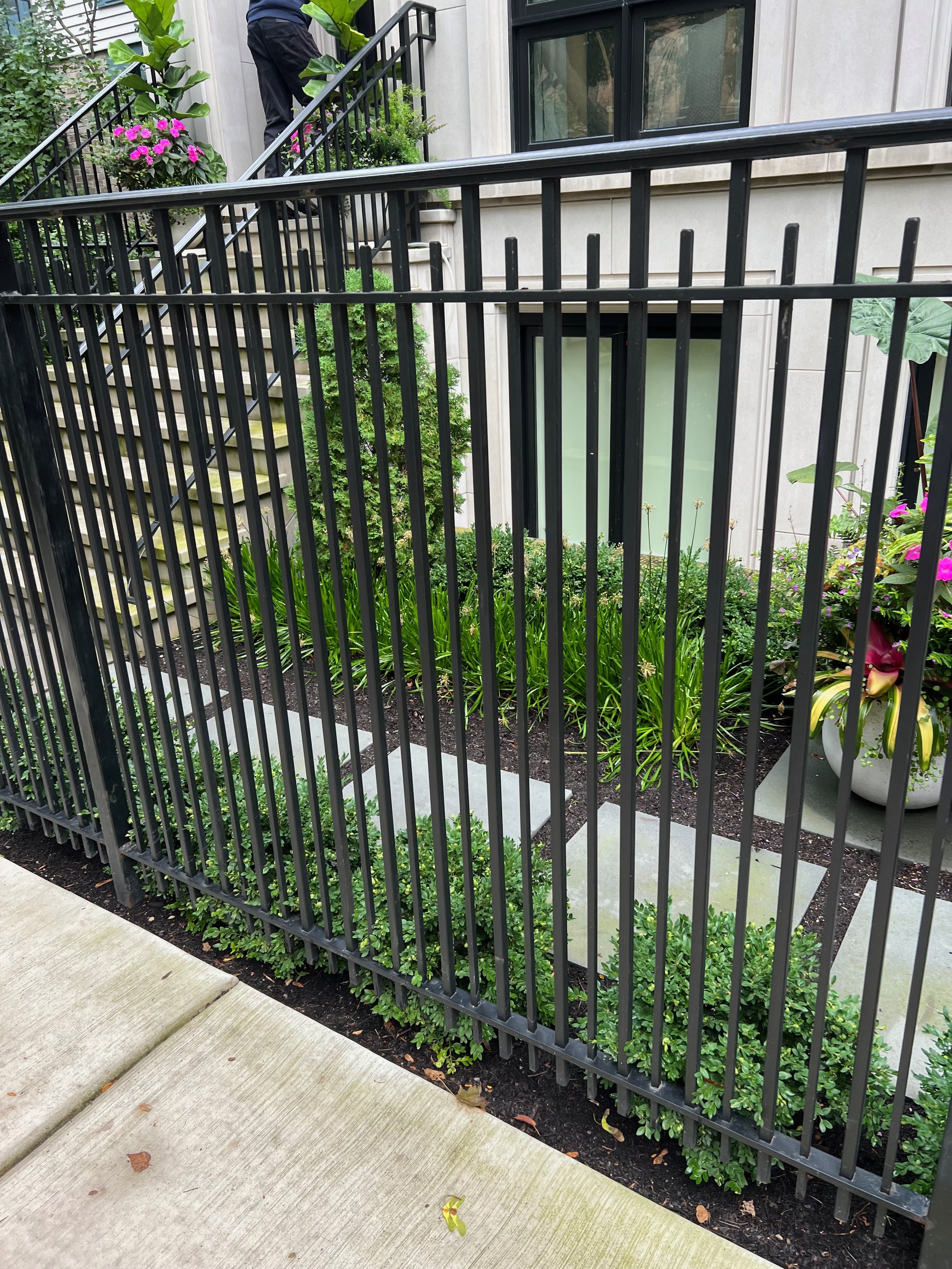 A black metal railing encircles a small garden area with green plants and colorful flowers in front of a beige building with large windows. A person is partially visible in the background on a staircase.