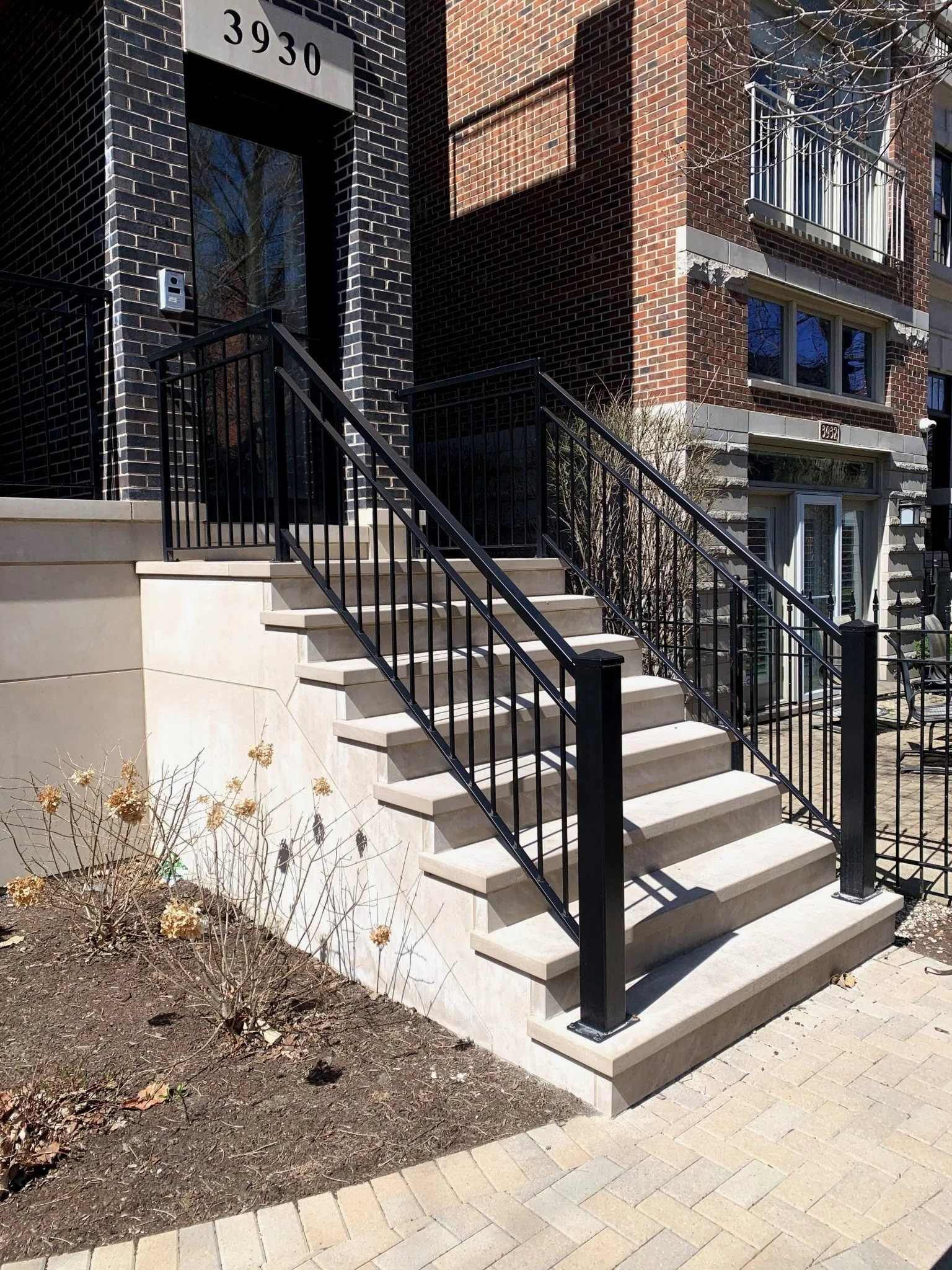 Exterior of a brick residential building with stairs leading up to the entrance, black metal railings, and a small garden with dried plants.
