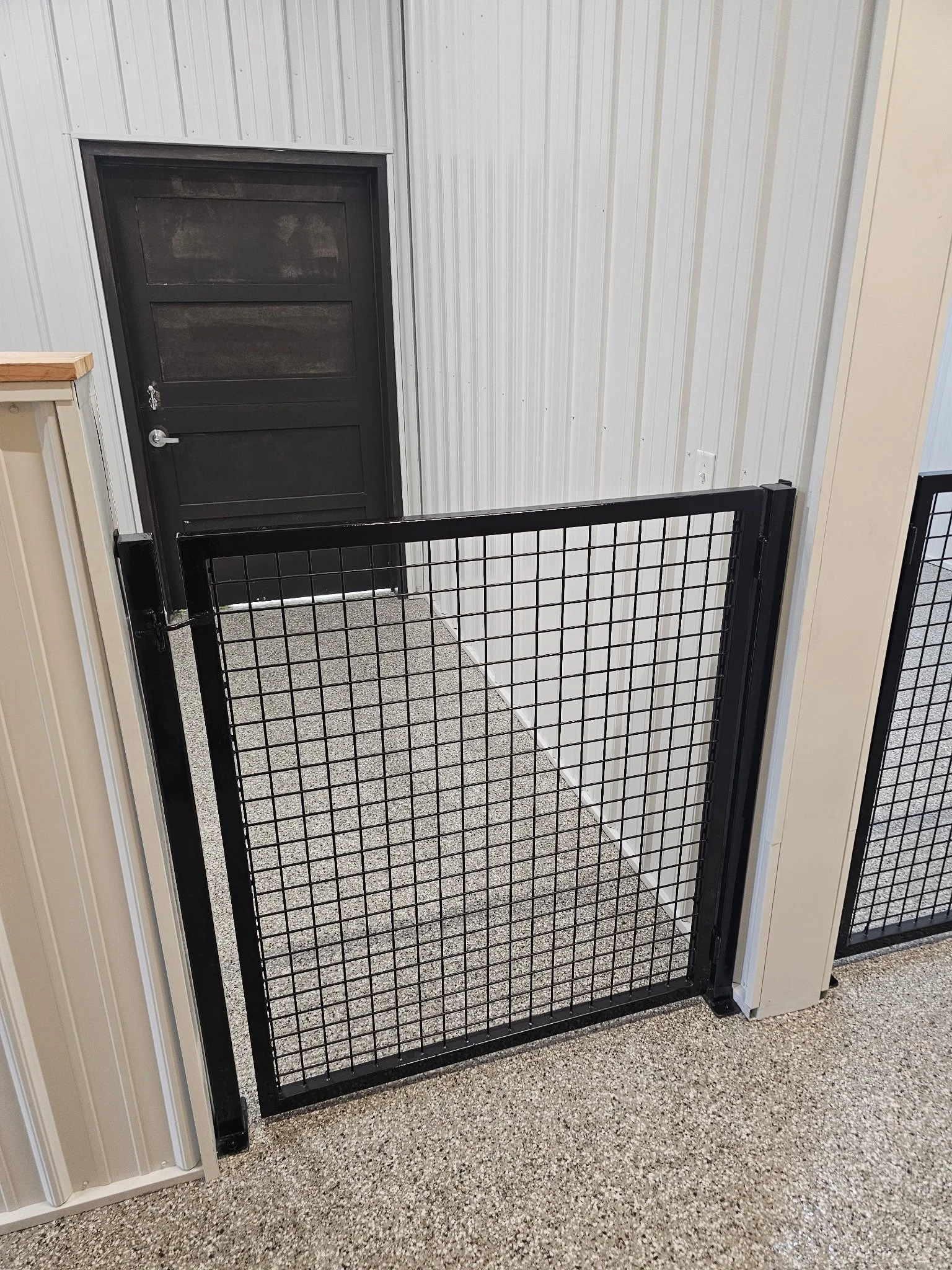 A black metal pet gate attached to a wall, blocking entrance to a door with a black frame and sliding panel, in an indoor setting with beige and cream-colored walls and a speckled beige carpeted floor.