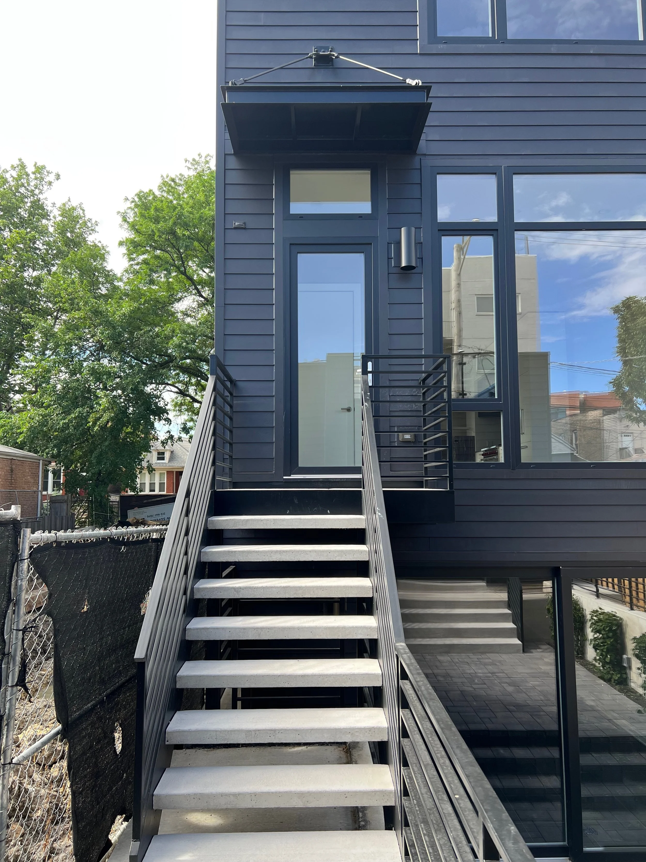 A modern black exterior building with a staircase leading to a door and large glass windows reflecting blue sky and clouds.