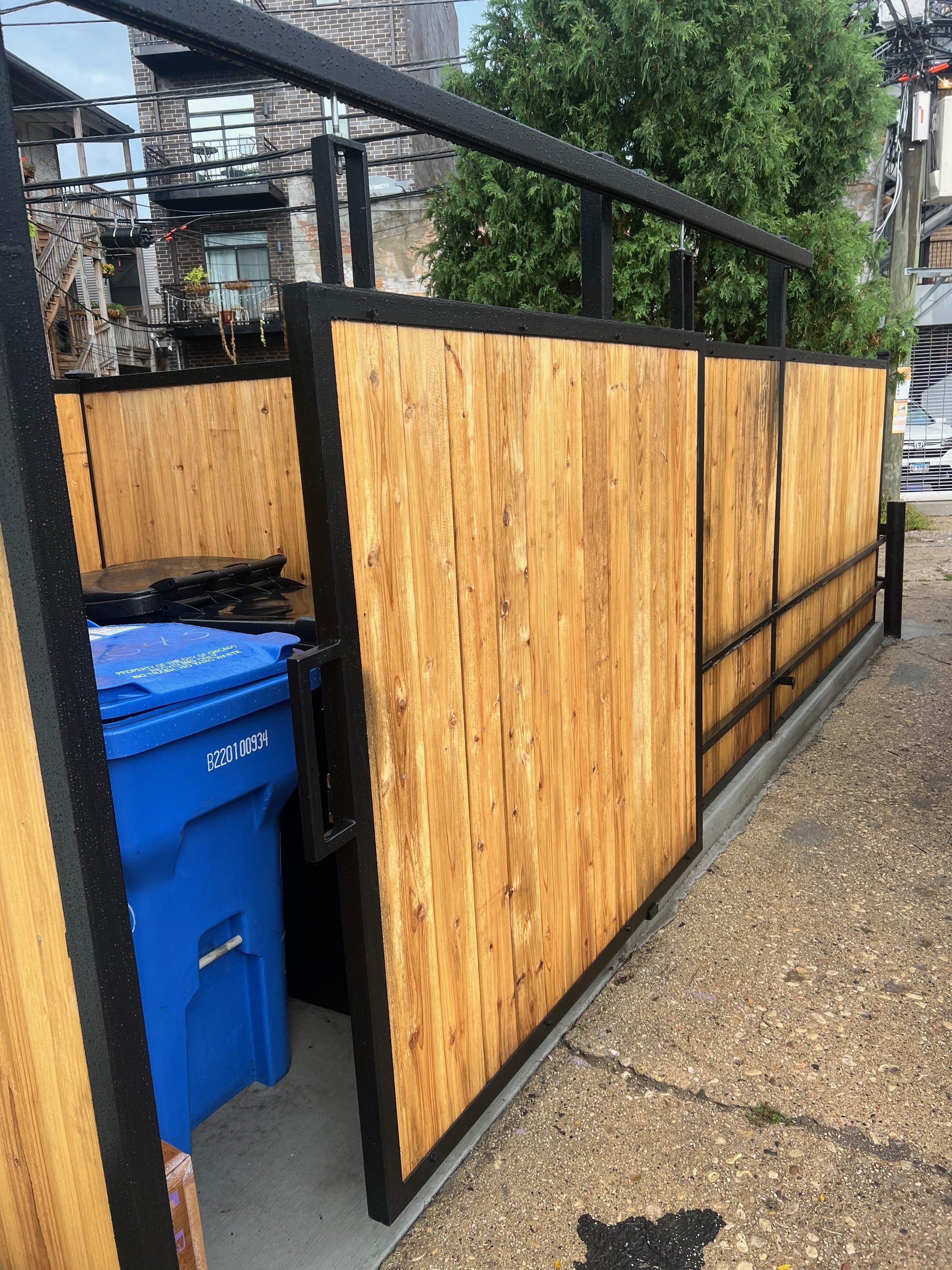 Wooden privacy fence with black metal framing surrounding a blue recycling bin on a concrete pad outside residential buildings and trees.