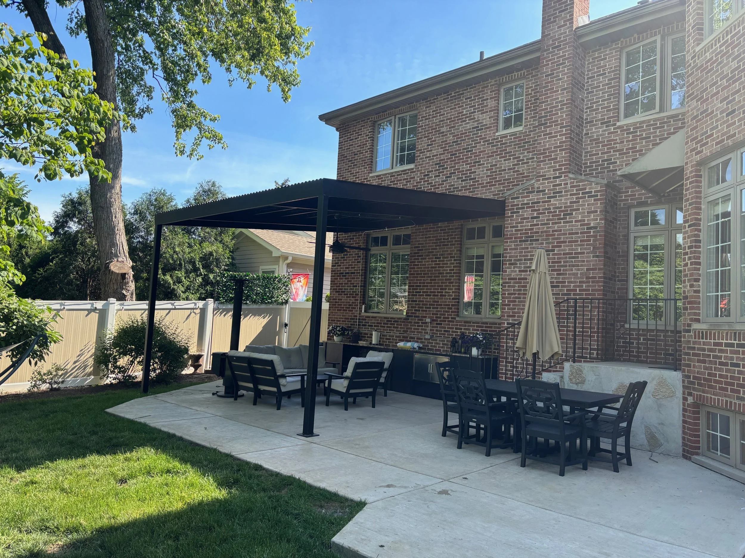 Backyard patio area with outdoor furniture, a covered seating area, a brick house with multiple windows, and a large tree with green leaves.