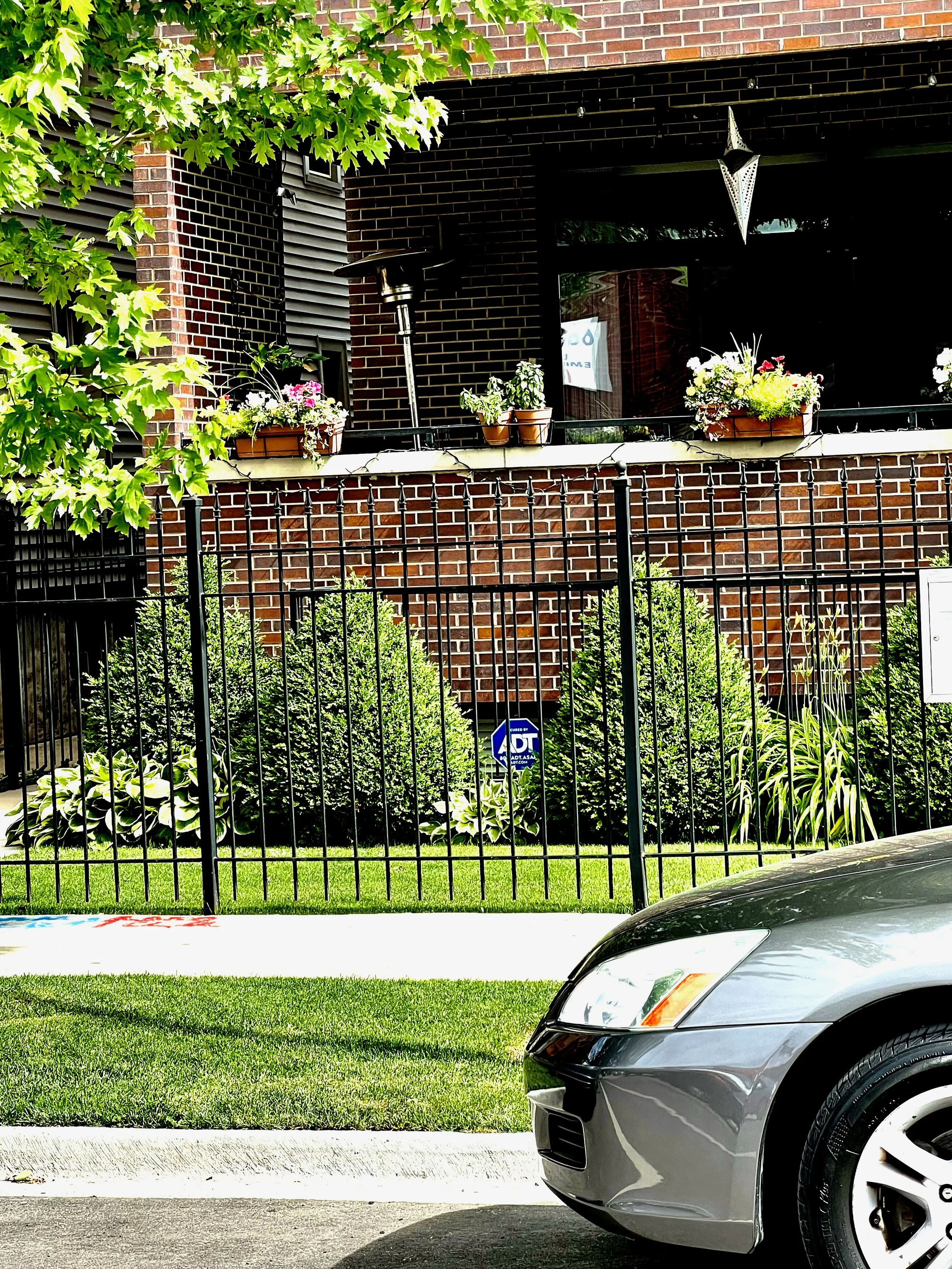 A gray car parked on a driveway in front of a front yard with green grass, bushes, and a black fence. Behind the yard, there is a building with a brick exterior, potted plants on the balcony, and a patio heater. There are also trees and bushes surrou