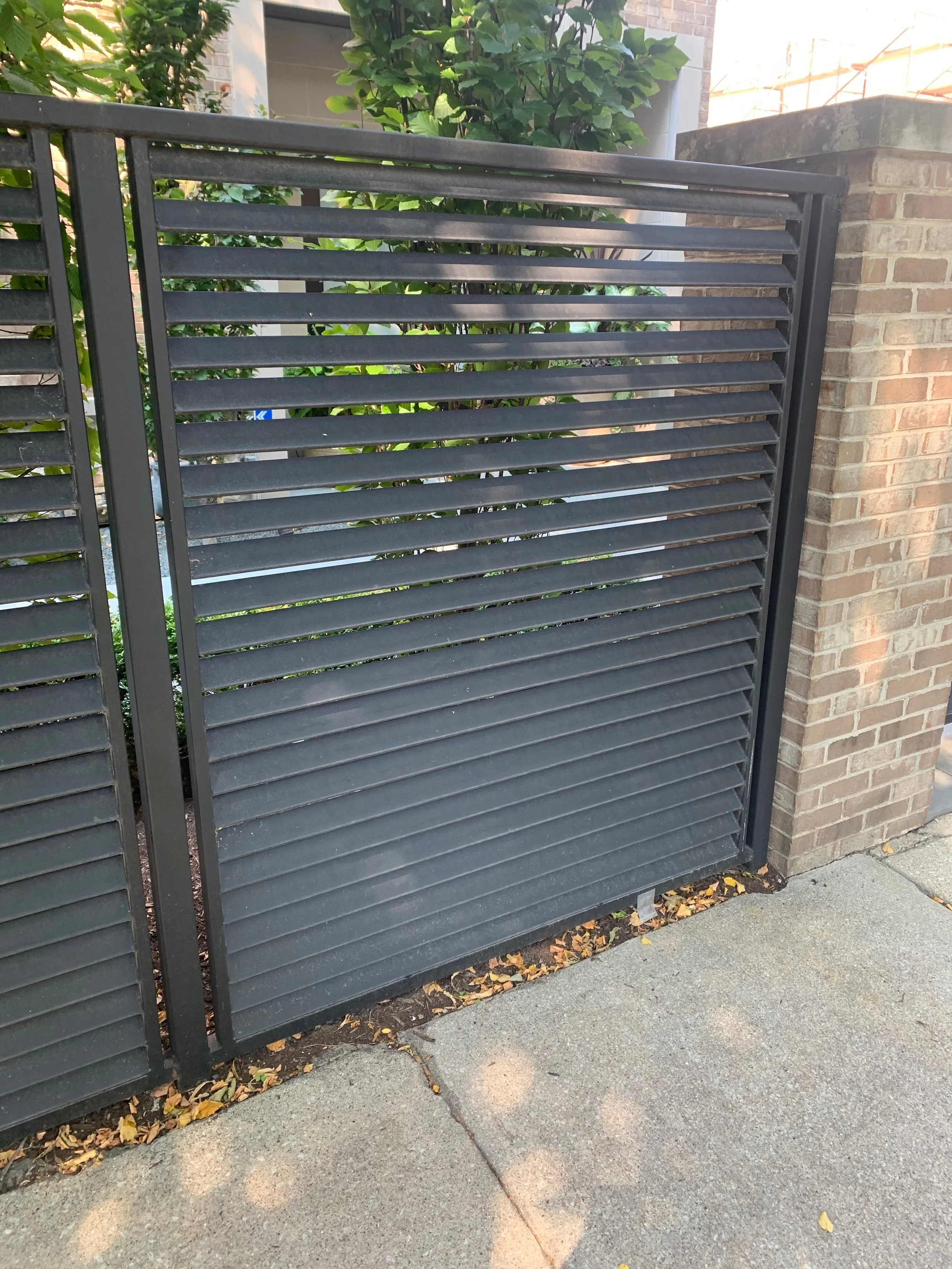 A black metal gate with horizontal slats, situated next to a brick wall with green foliage in the background, and a sidewalk with scattered fallen leaves in the foreground.