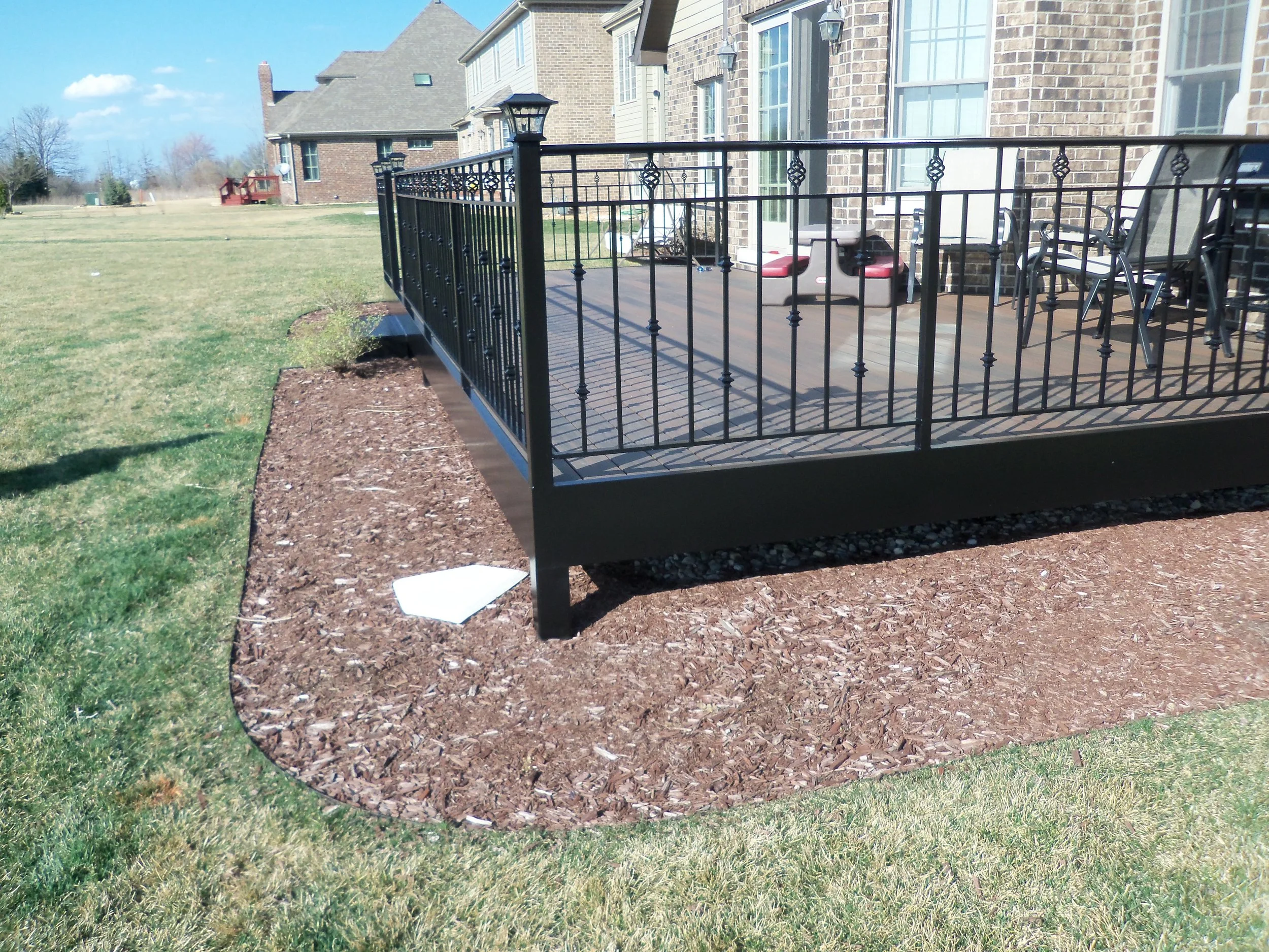 A backyard patio with a black metal railing, outdoor chairs, and a small table. There are brick houses in the background and a clear blue sky.