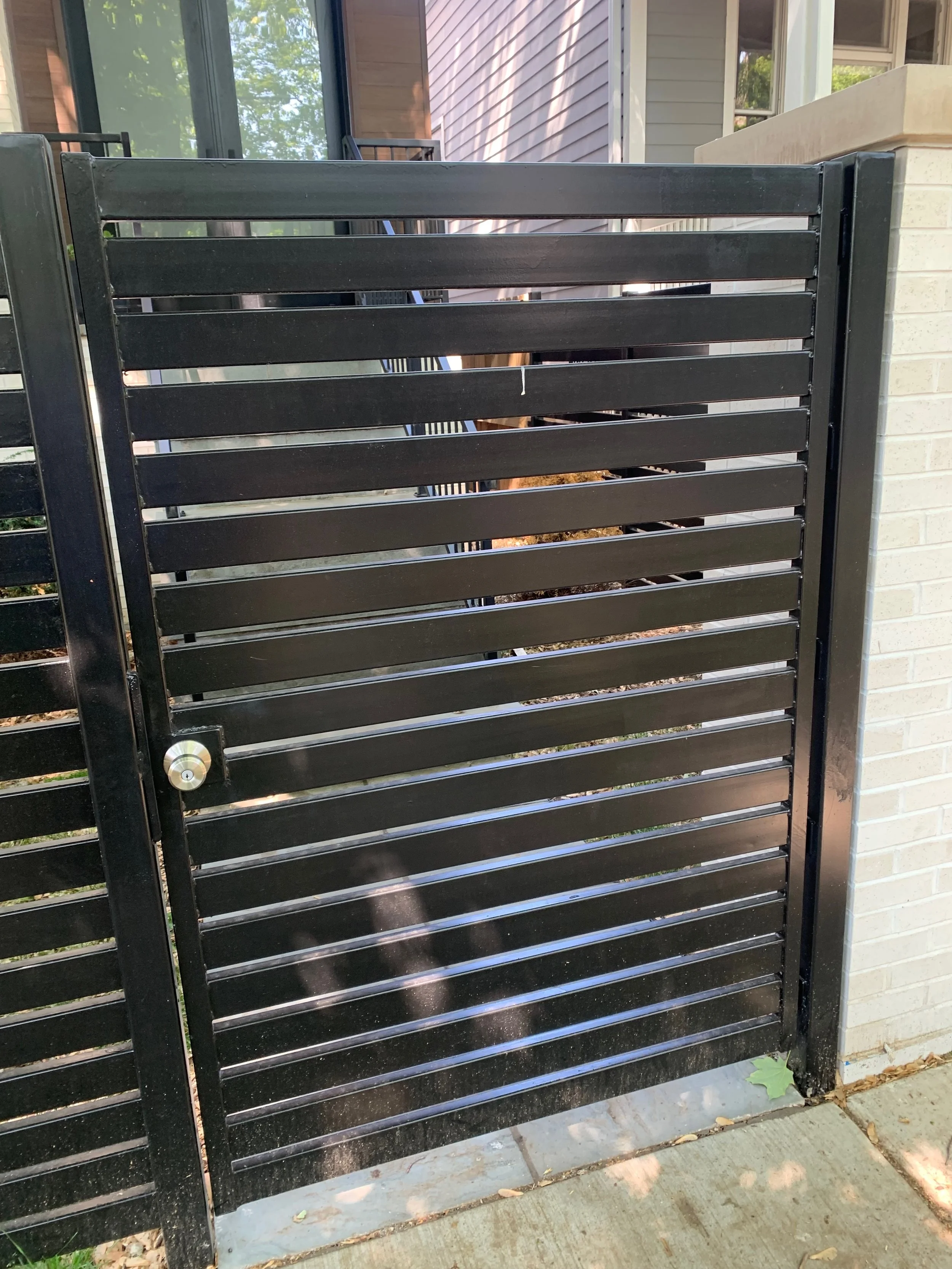 A black metal gate with horizontal slats situated in front of a house with siding, a window, and a brick column.