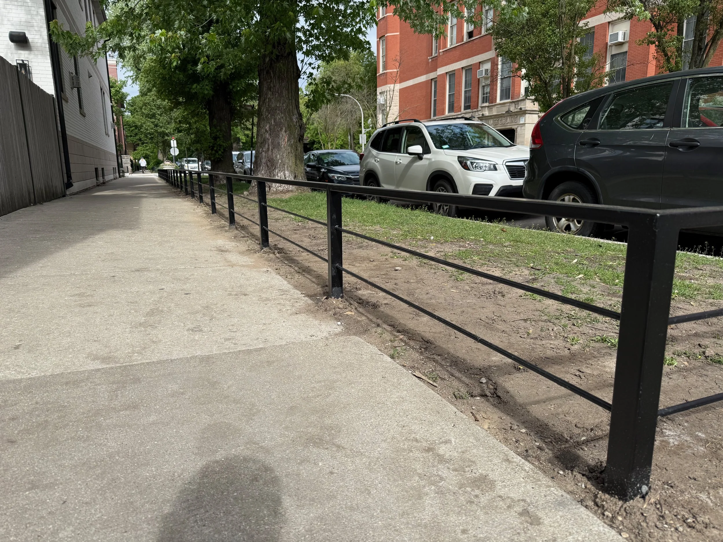 Sidewalk with trees and parked cars, including a white SUV and a black vehicle, in an urban area with residential buildings in the background.