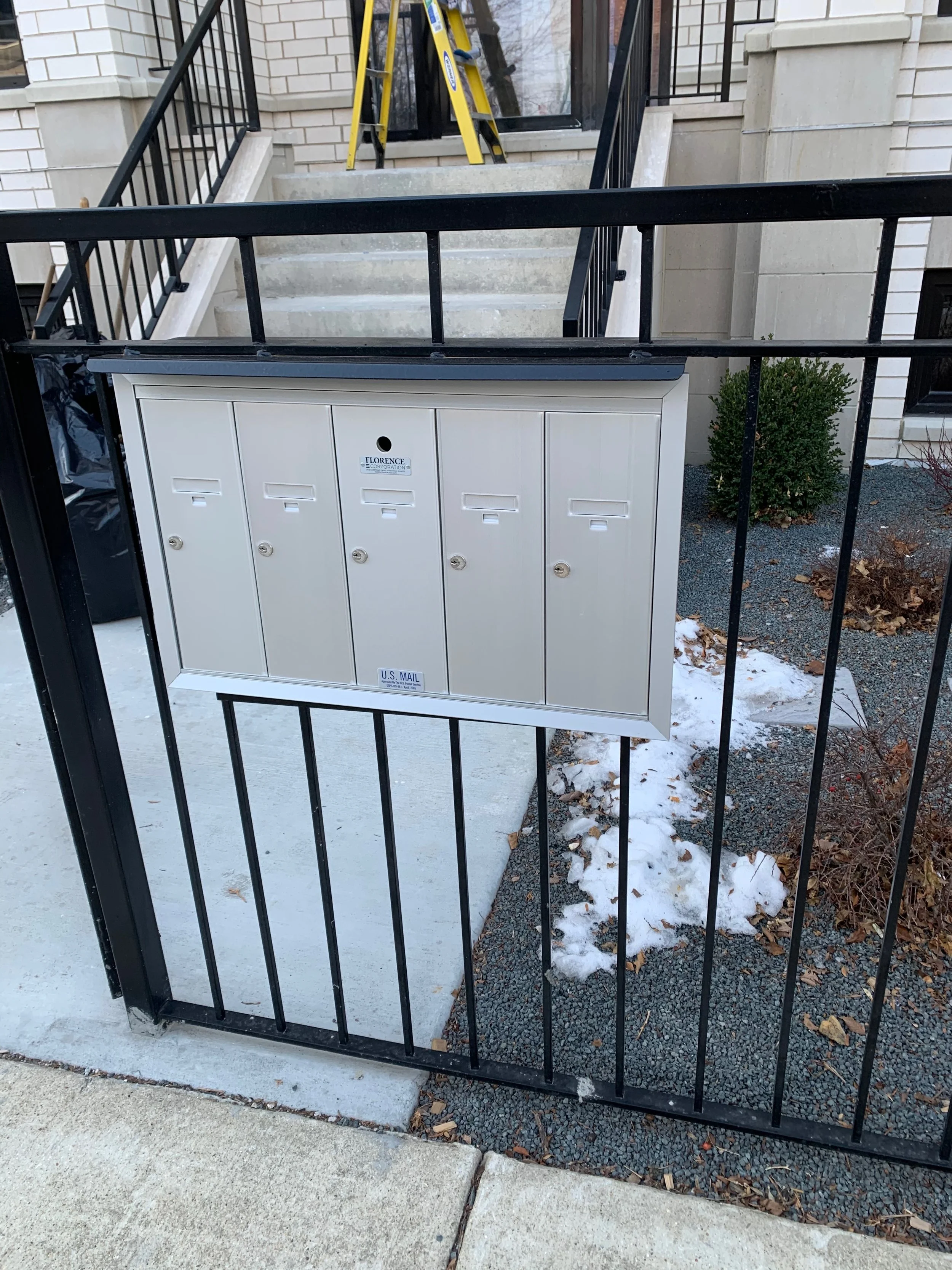 Front yard of a house with a black metal fence, concrete walkway, snowy ground, small green bushes, and a white mailbox box labeled 'U.S. Mail' attached to the fence.
