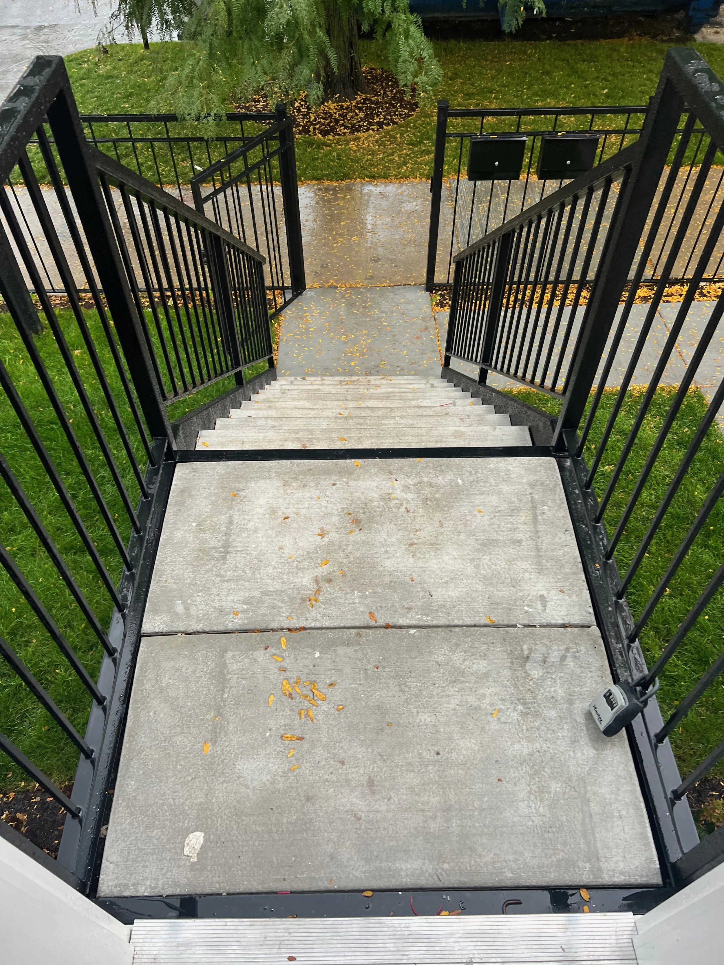View from the top of a staircase leading down to a sidewalk. The staircase has black metal railings on both sides and concrete steps. The sidewalk is wet with fallen yellow leaves scattered across. There is a grassy area with a tree and some bushes n