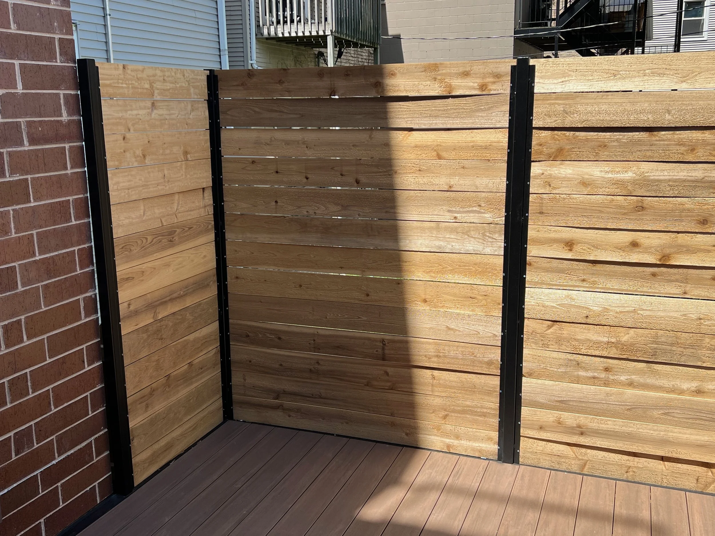 Wooden privacy fence with black metal posts on an outdoor balcony or patio area, with part of a brick wall and neighboring houses in the background.