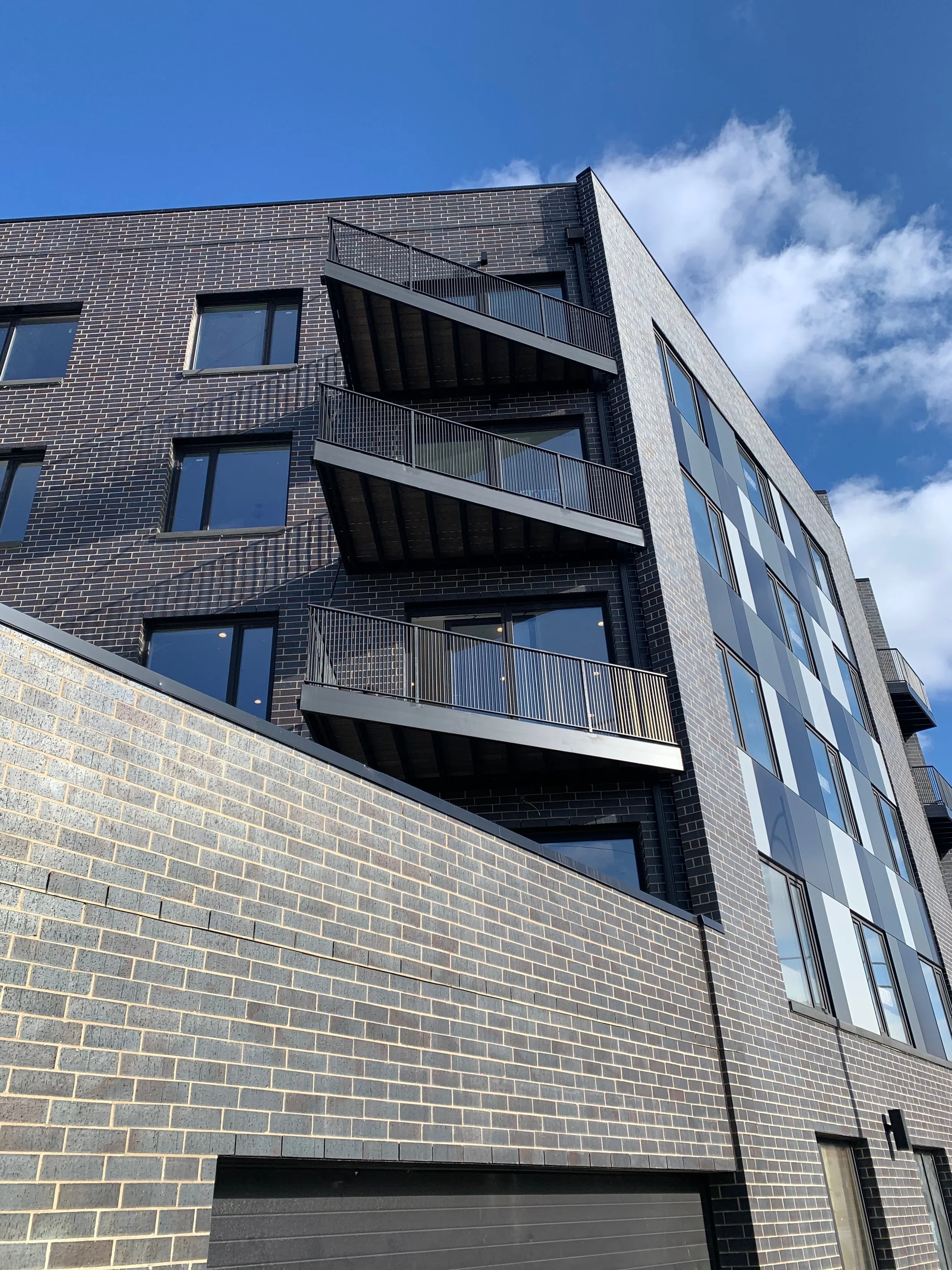 Modern multi-story building with dark brick facade, black metal balconies, large windows, and a clear blue sky with some clouds.